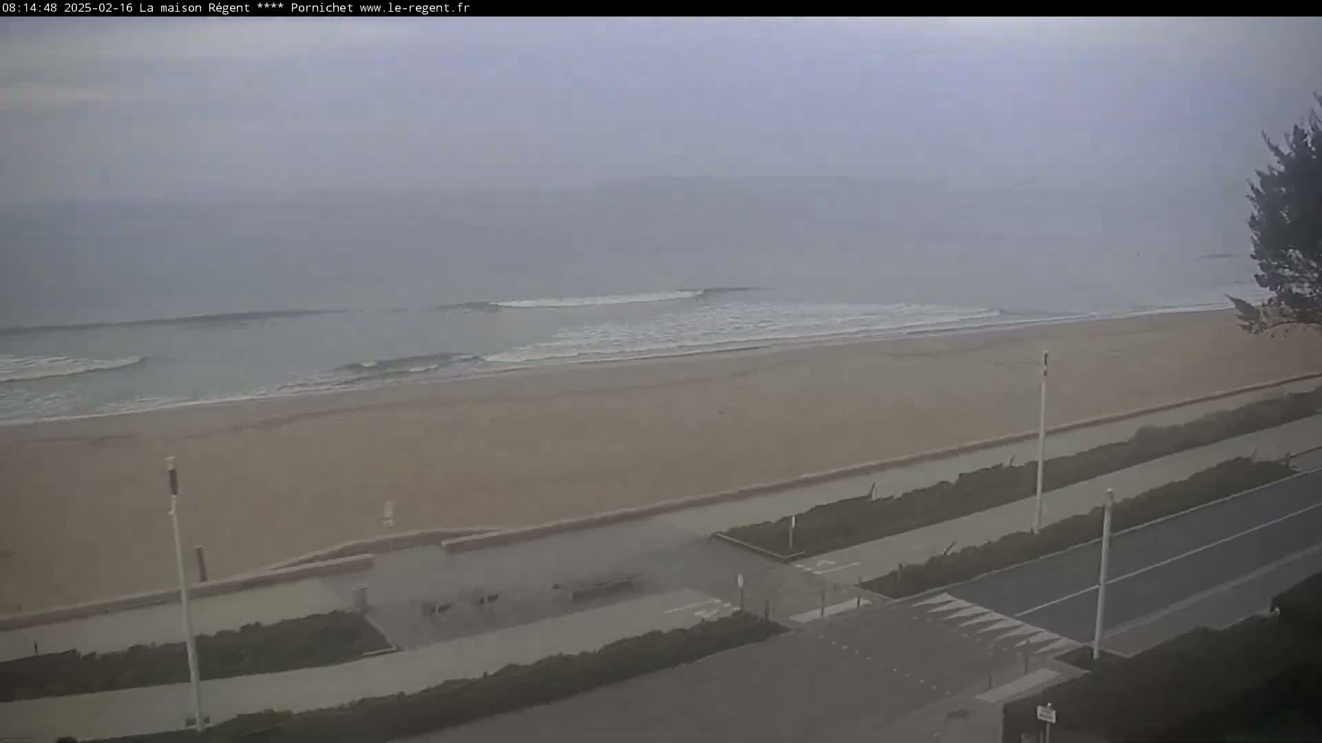 A long, sandy beach with small waves under an overcast sky is viewed from above, showing a paved road and pedestrian path leading away from the shore.