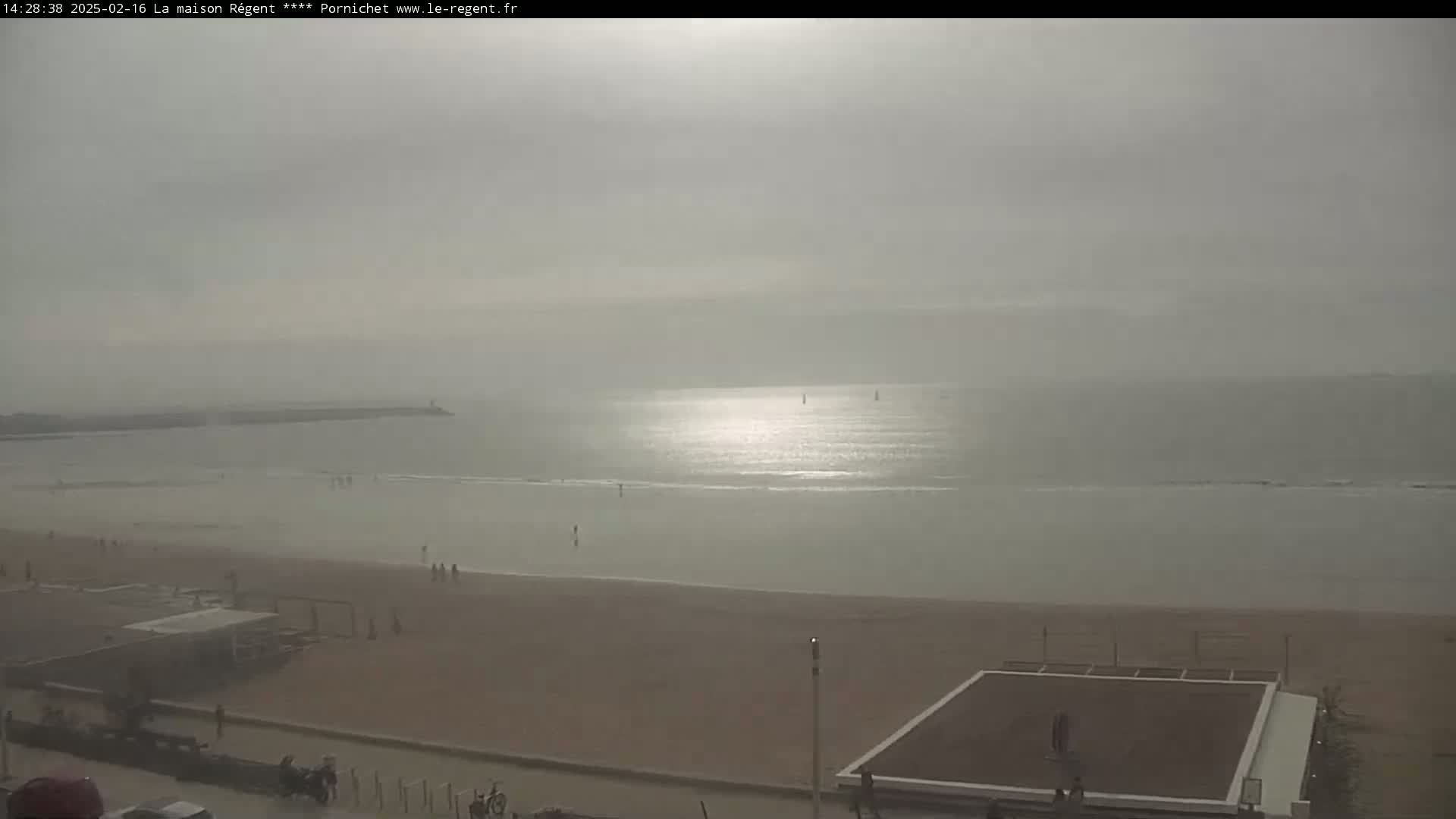 A hazy beach scene shows a calm sea under a mostly overcast sky, with a few people on the sand and a structure resembling a beach volleyball court.
