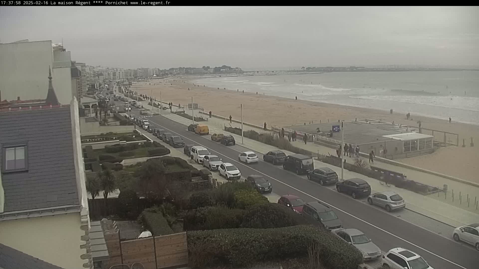 A coastal road with parked cars lines a sandy beach with a calm ocean under an overcast sky.