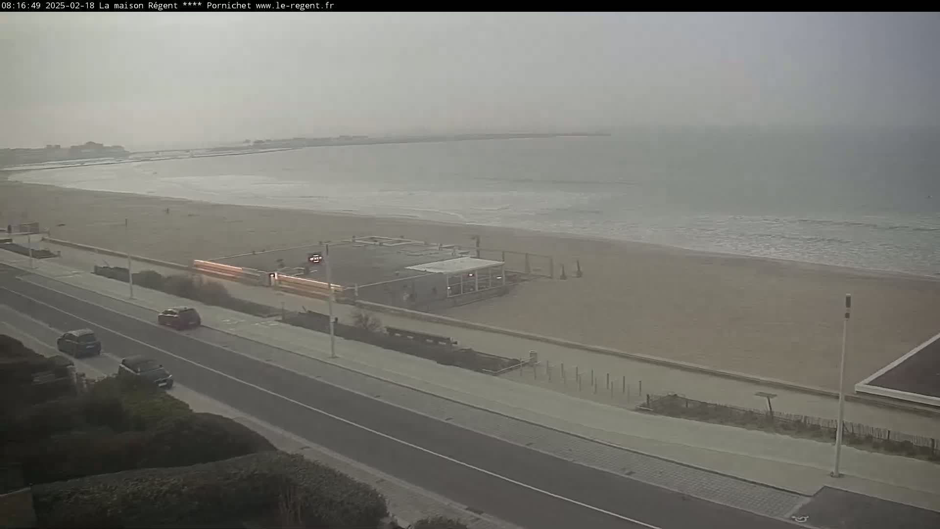 A hazy view shows a beach with calm waves, a road with a few cars, and a building near the shore.