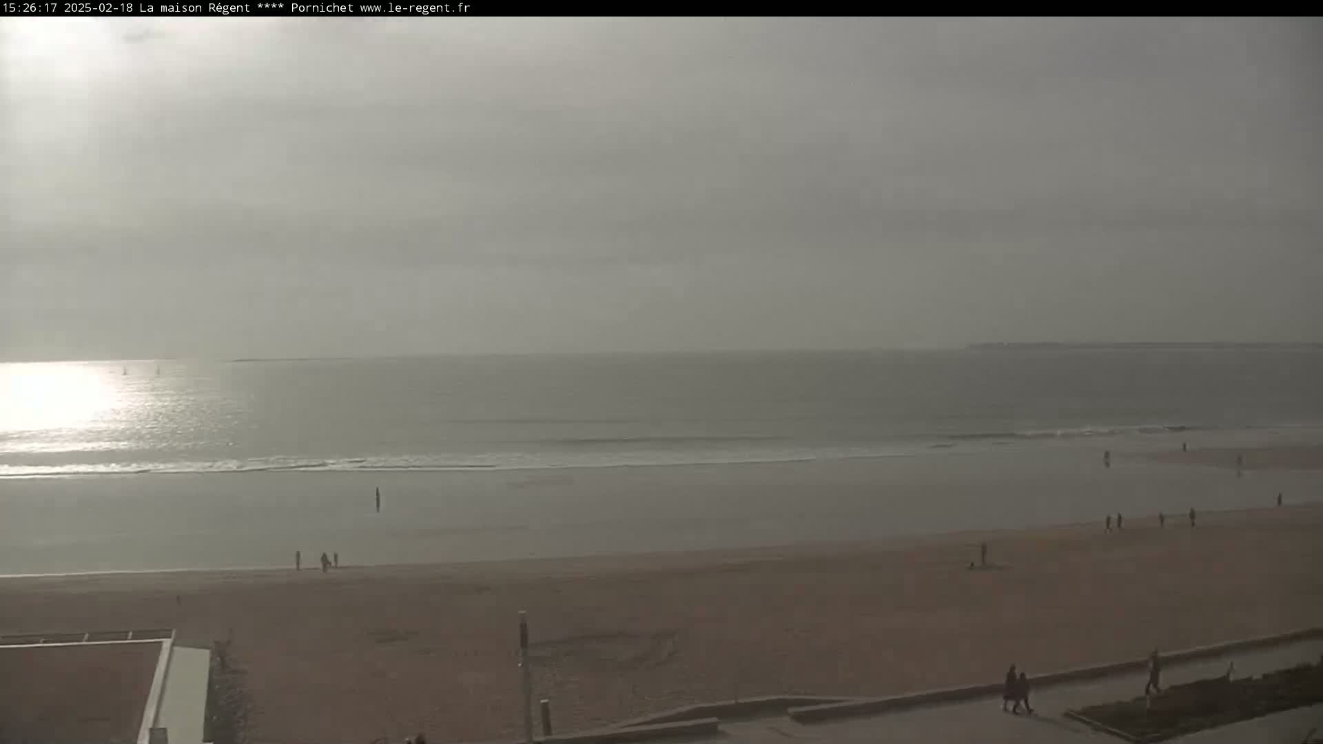 A mostly overcast day reveals a wide sandy beach with a few people strolling, leading to a calm ocean with gentle waves under a hazy sky.