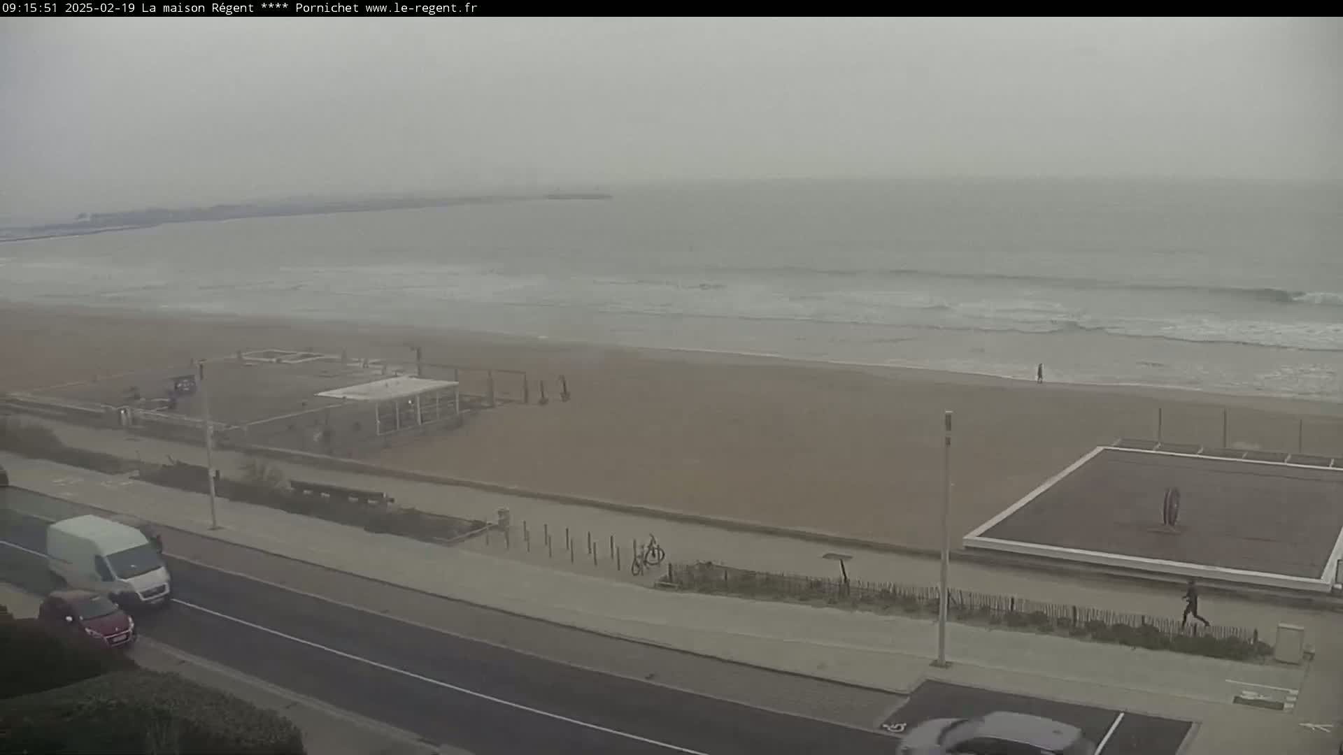 A hazy, overcast day shows a beach with a few people, a road with cars, and outdoor exercise equipment near the ocean.