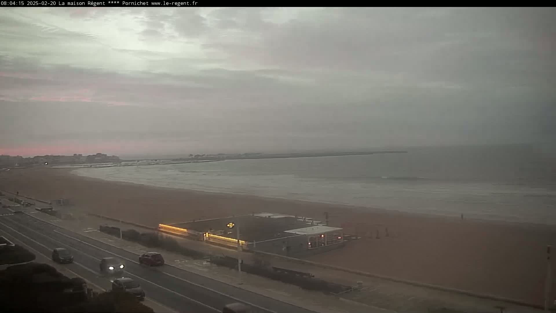 An overcast sky hangs over a beach with calm waves, a road with cars, and a building near the shore.