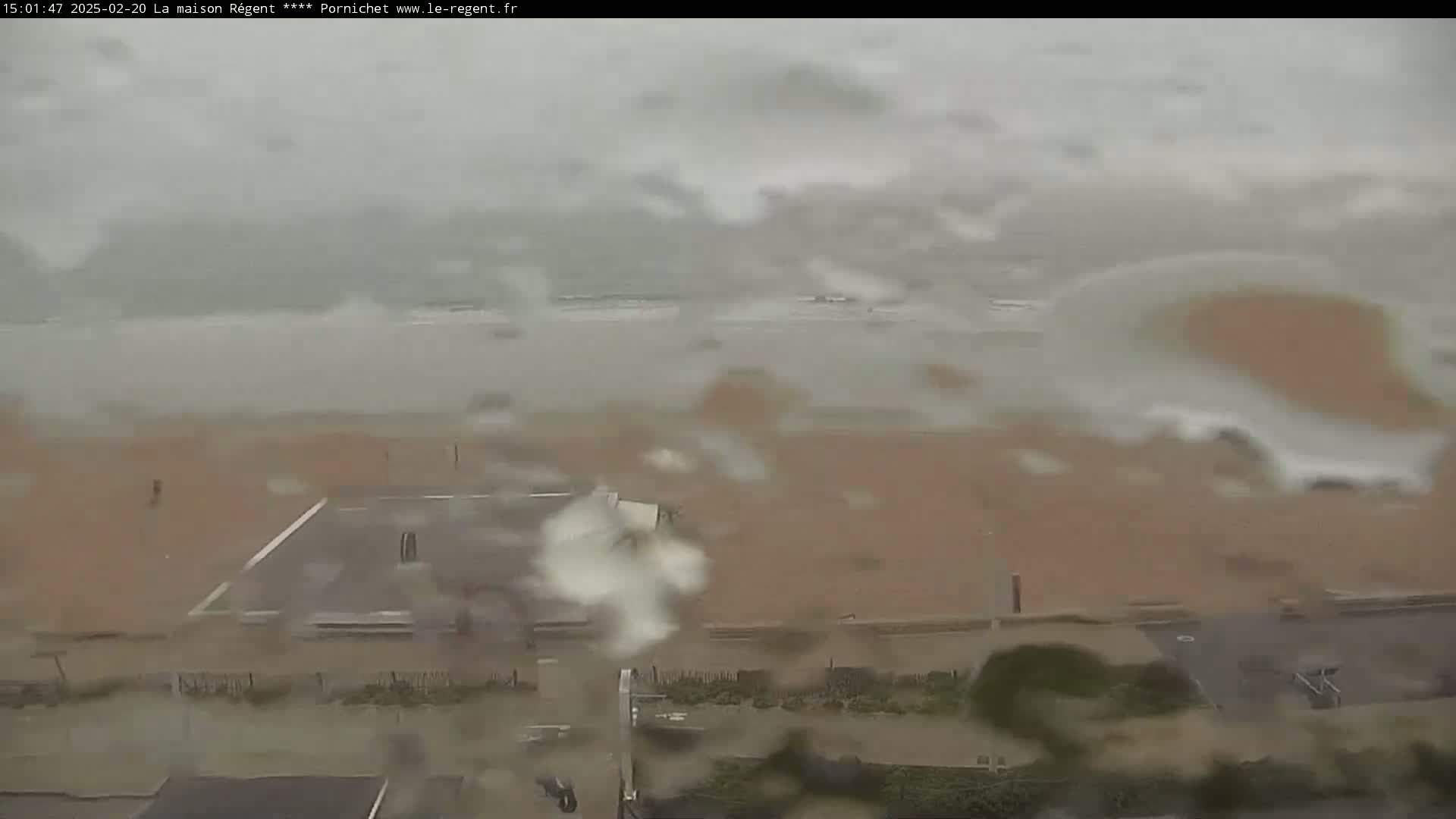 A rainy view of a beach with an overcast sky shows waves crashing on the shore and a mostly empty sandy beach, with a parking lot and some vegetation visible in the foreground.
