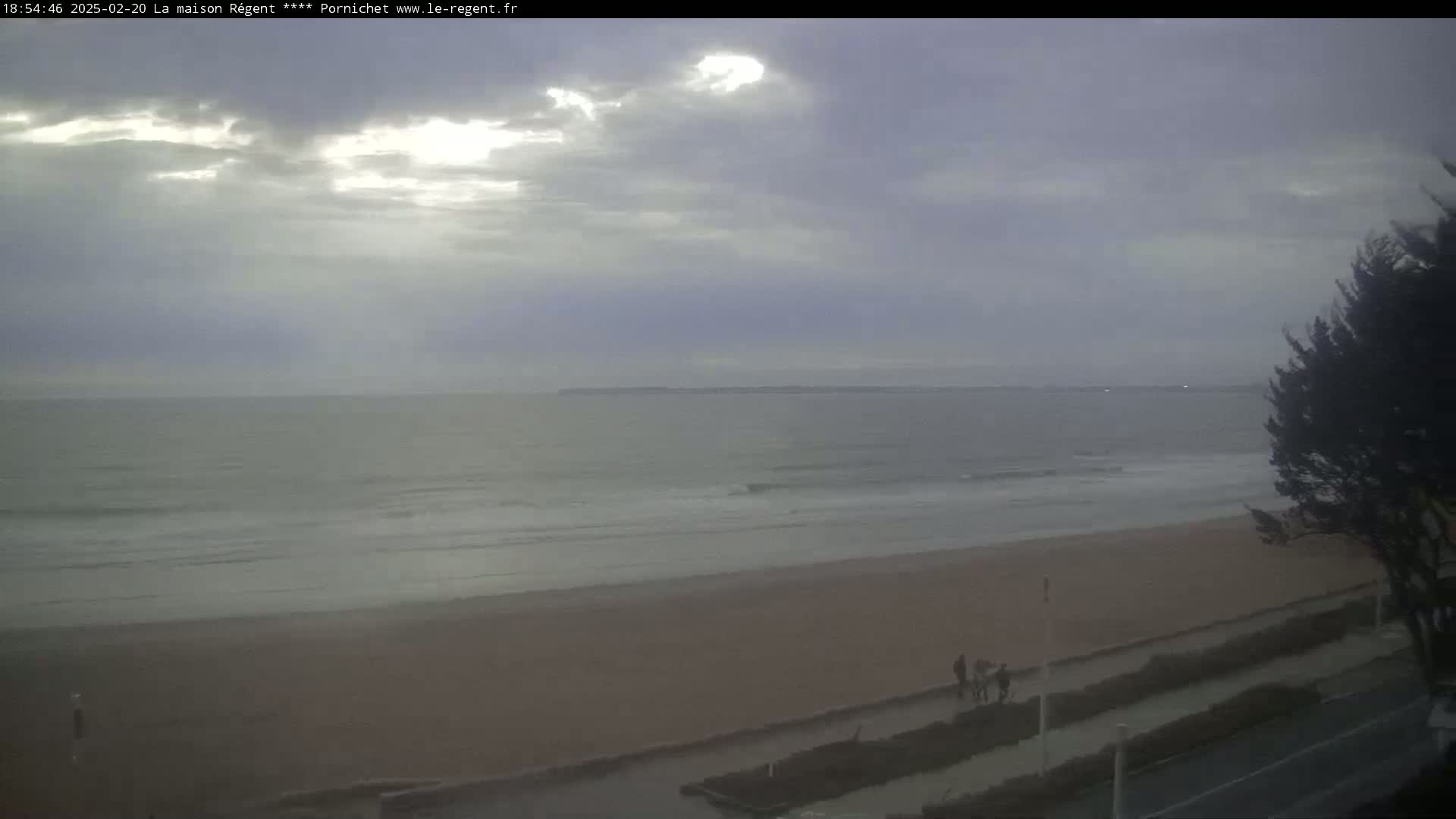 A mostly cloudy day reveals a wide sandy beach and calm ocean waves, with a few people walking along the shore.