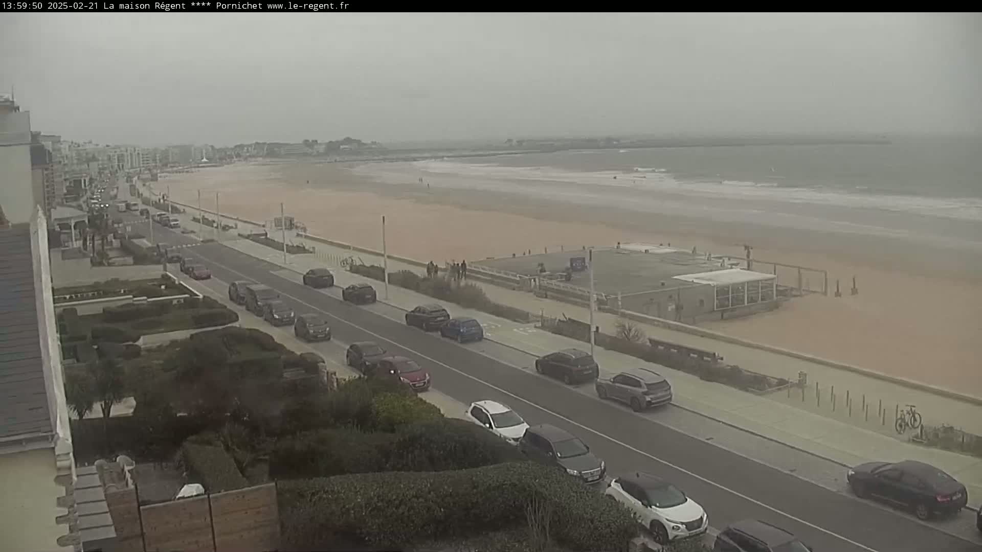 A hazy, overcast day shows a beach with a few people, a road lined with parked cars, and buildings in the distance.