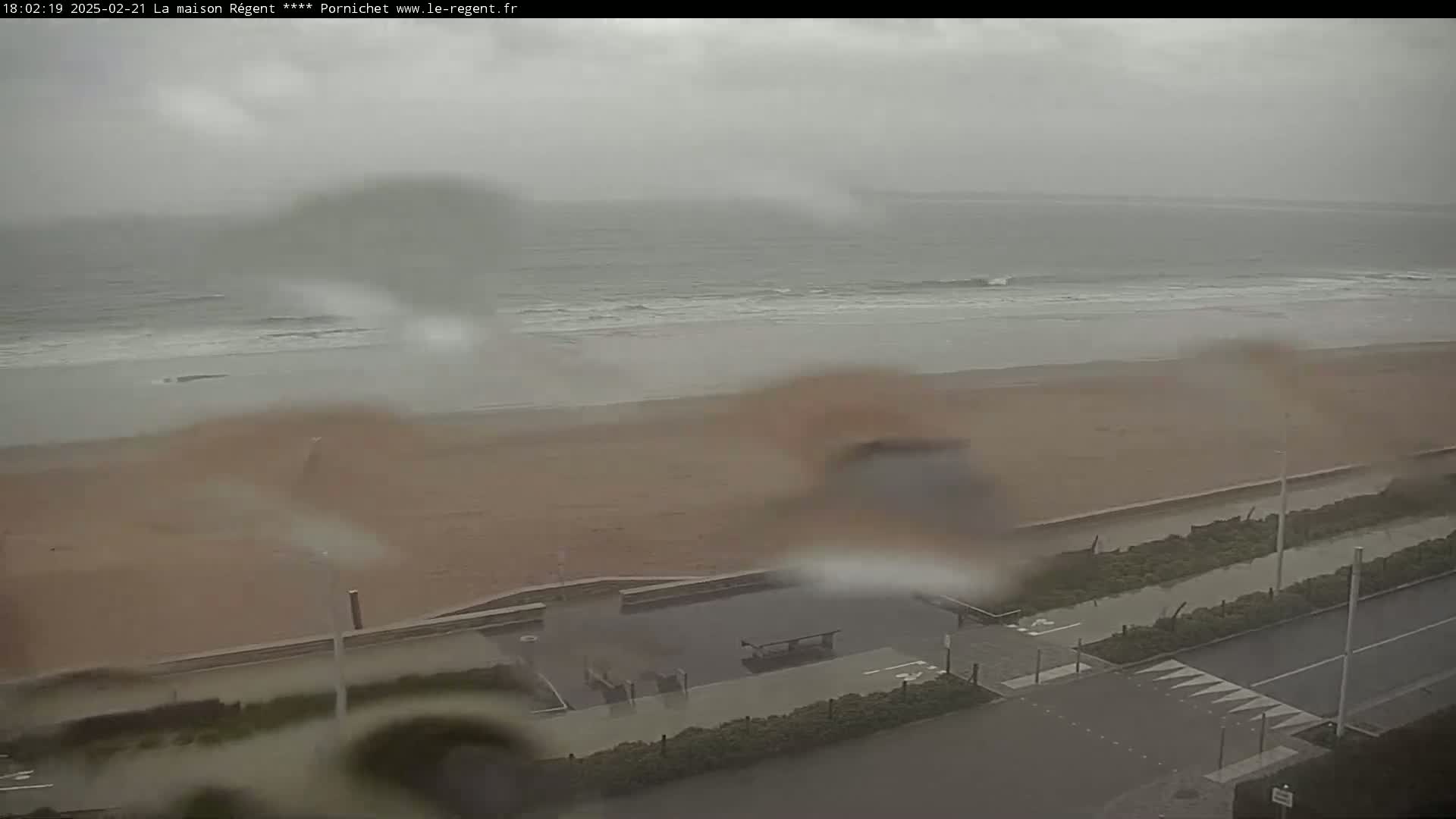 A rainy view of a beach with a road and some vegetation bordering it, showing a mostly grey ocean with small waves.