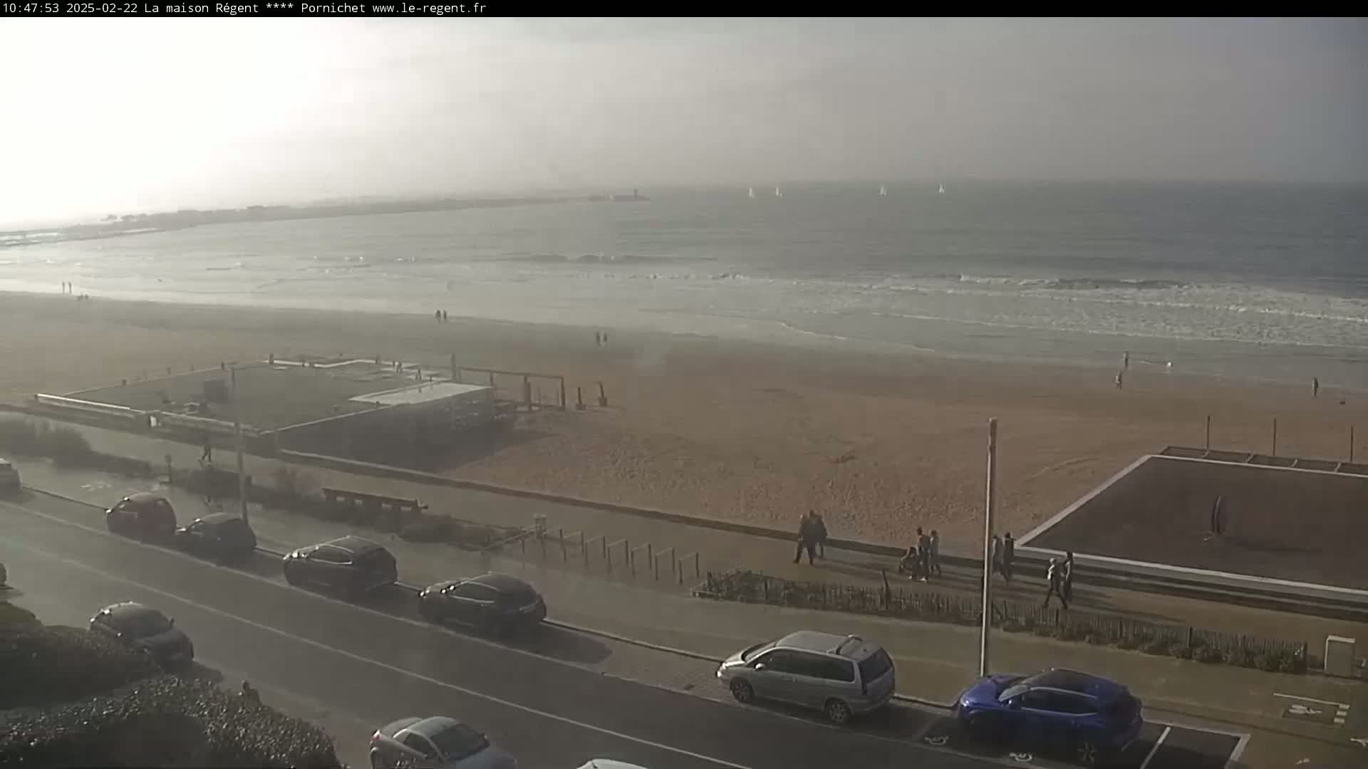 A hazy, overcast day shows a sandy beach with several people walking along the shore and parked cars lining a road overlooking the ocean where a few sailboats are visible.