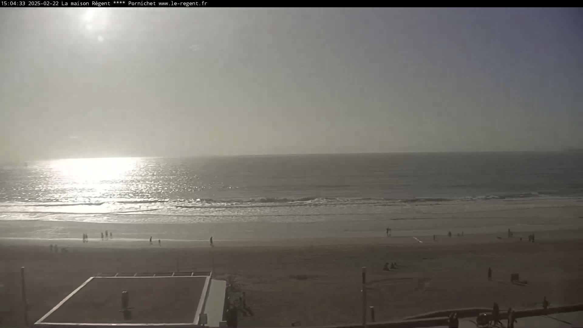 A hazy, sunny day at a beach shows a calm ocean with small waves, a sandy shore with scattered people, and a flat-roofed structure near the bottom.