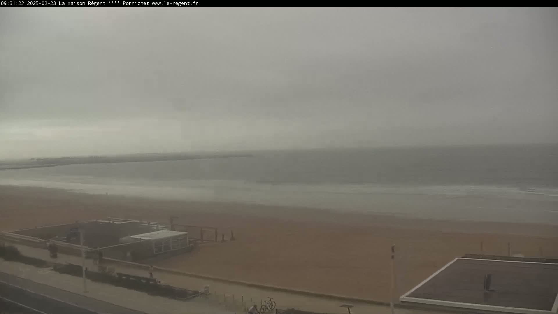 A hazy, overcast day shows a wide sandy beach and calm ocean with a few small structures near the shore.