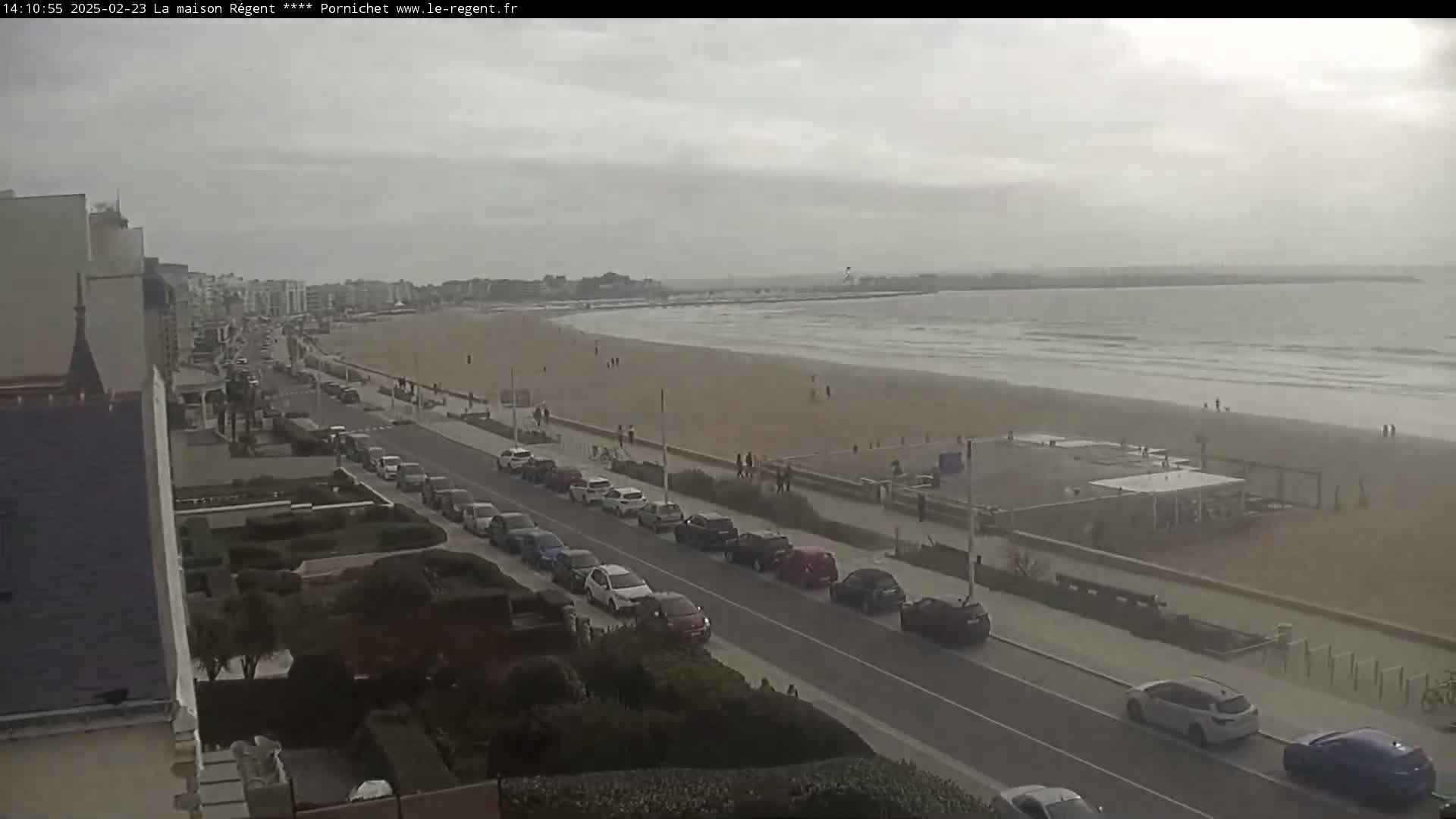 A mostly cloudy day shows a beach with a few people, cars parked along a road beside it, and buildings in the background.