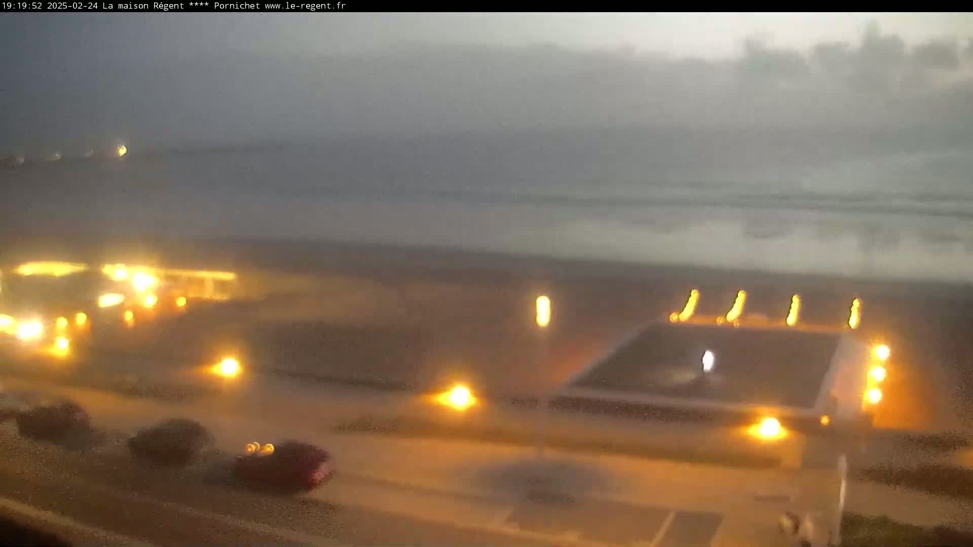 A hazy, dimly lit coastal scene at night shows a body of water, a beach with lights, and vehicles parked nearby.