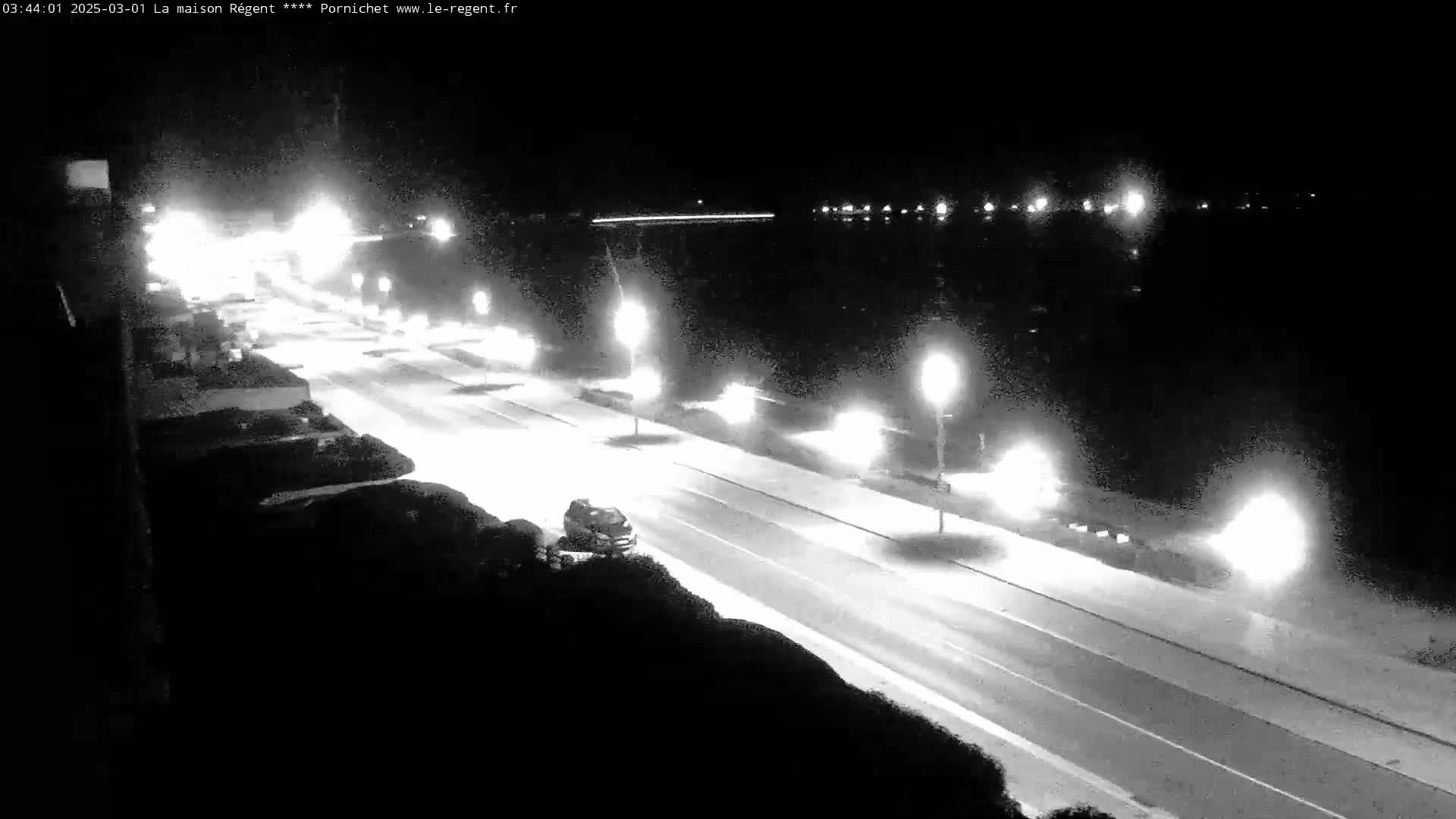 A nighttime, brightly lit roadway with a few vehicles is seen from an elevated vantage point, overlooking a body of dark water in the background.