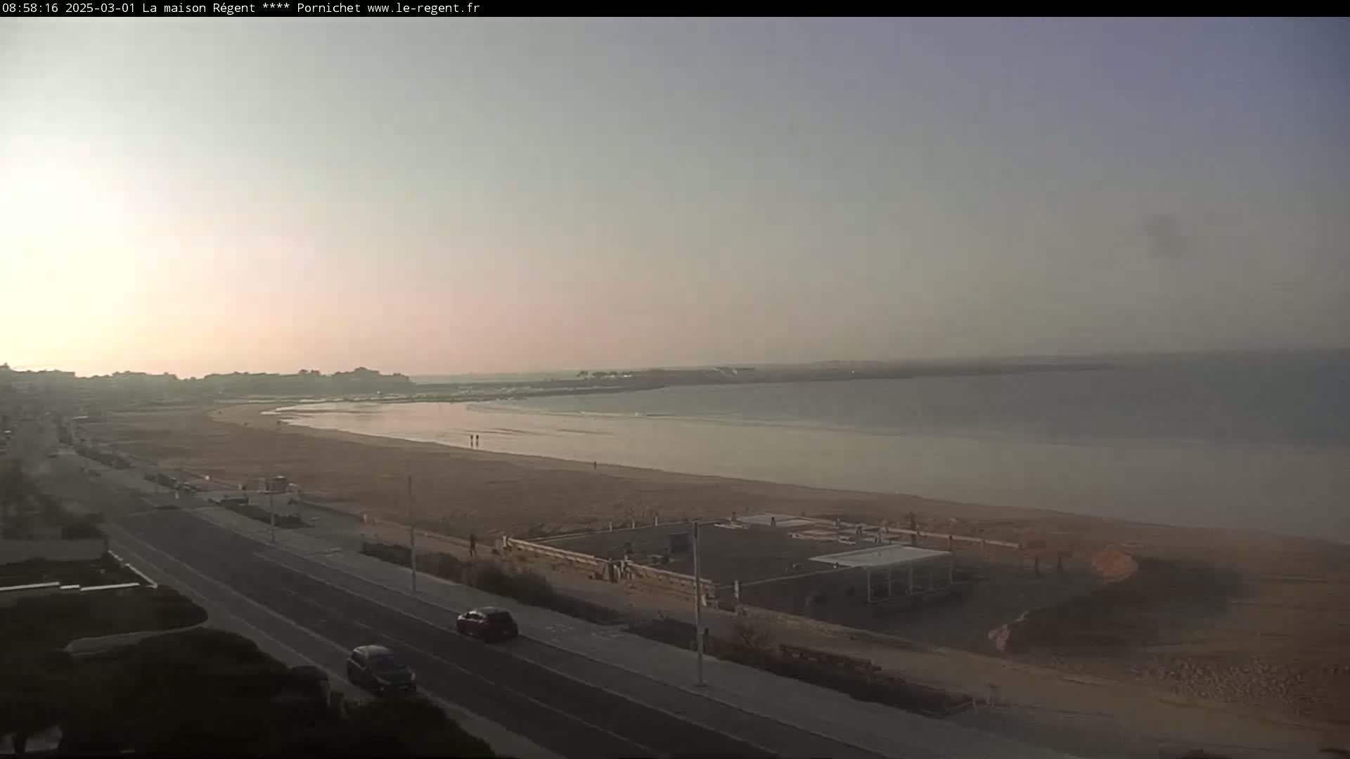 A long sandy beach stretches along a calm, pale-blue ocean under a hazy, light-pink sky.