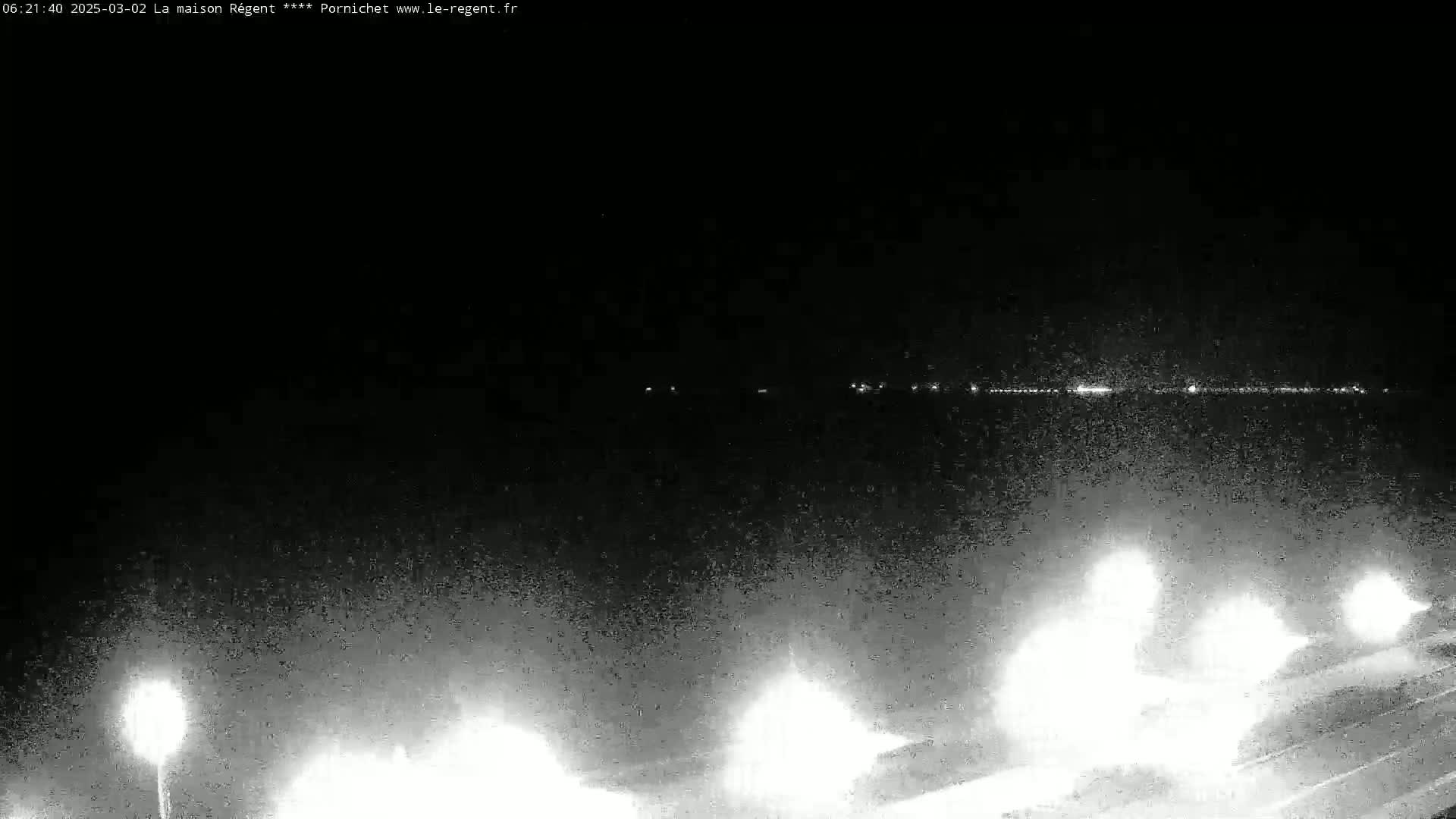 A nighttime, low-light view shows a dark body of water with a distant line of lights, and several bright lights illuminating the foreground from what appears to be a road or walkway.