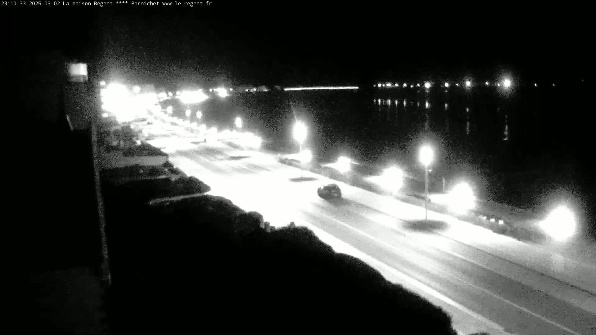 A nighttime, snow-covered road with cars driving on it, next to a body of water under a dark sky.