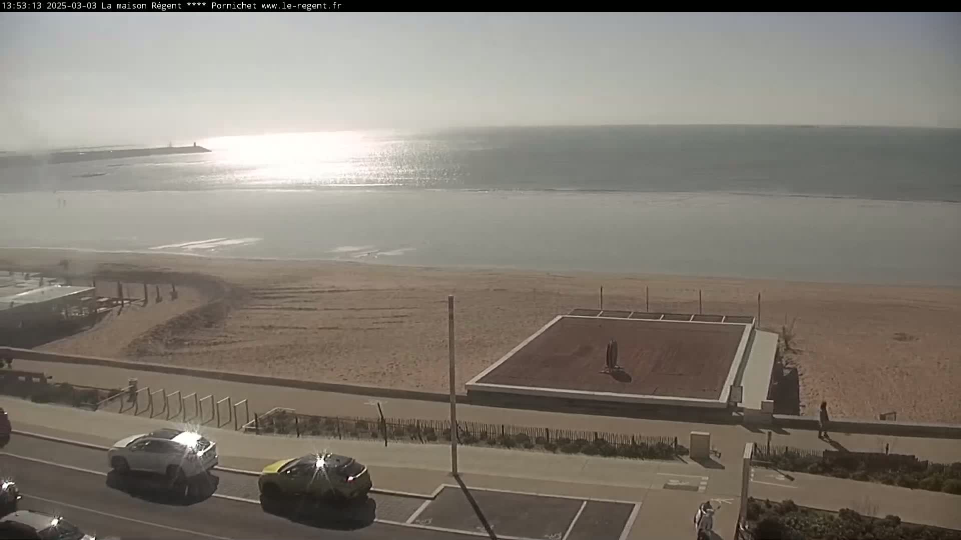 A sandy beach with a volleyball court and cars parked along a road overlooks a calm, sunlit ocean on a hazy day.