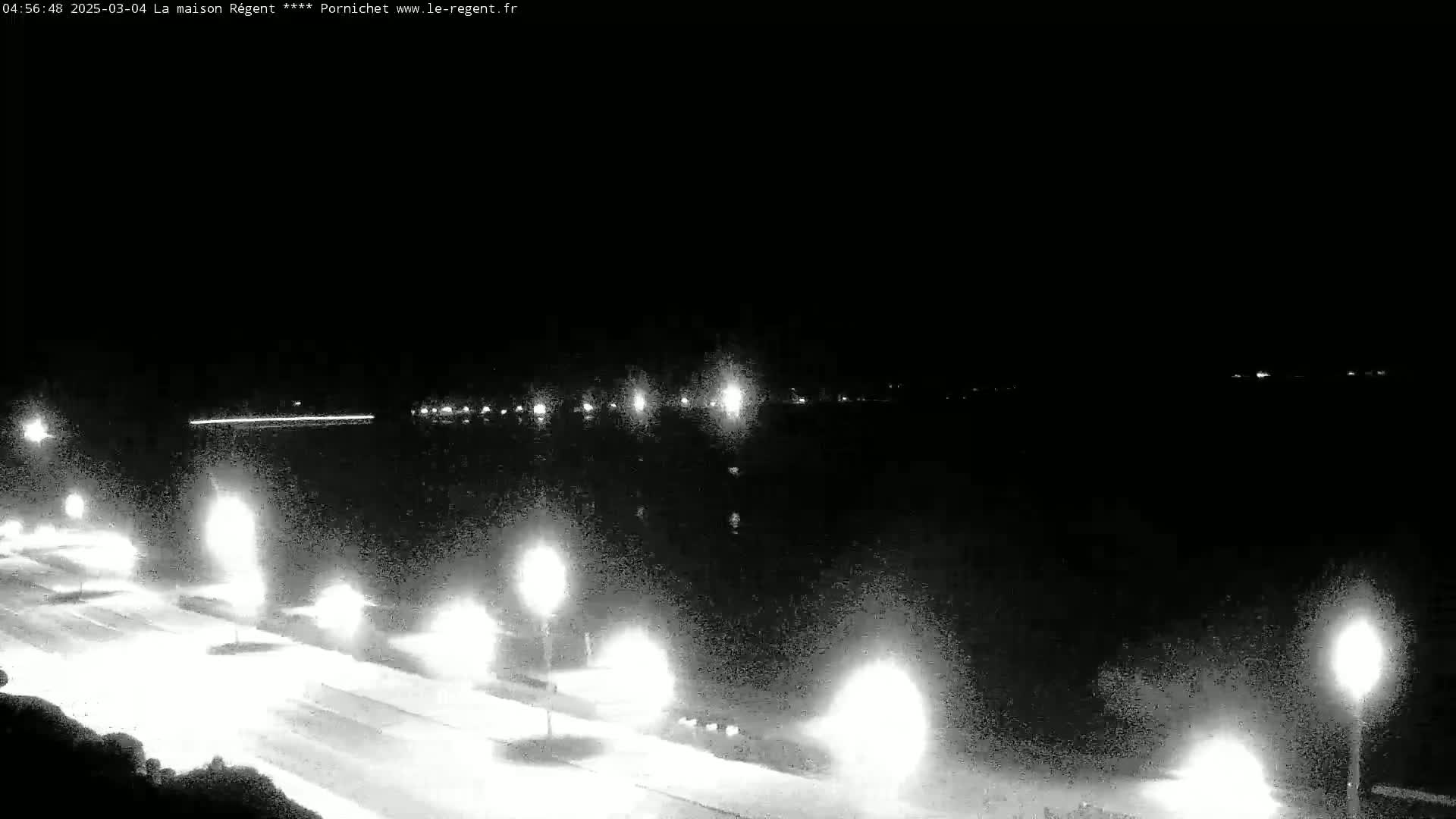 A nighttime, low-light view of a roadway lit by streetlights alongside a body of water with distant lights visible across the water.
