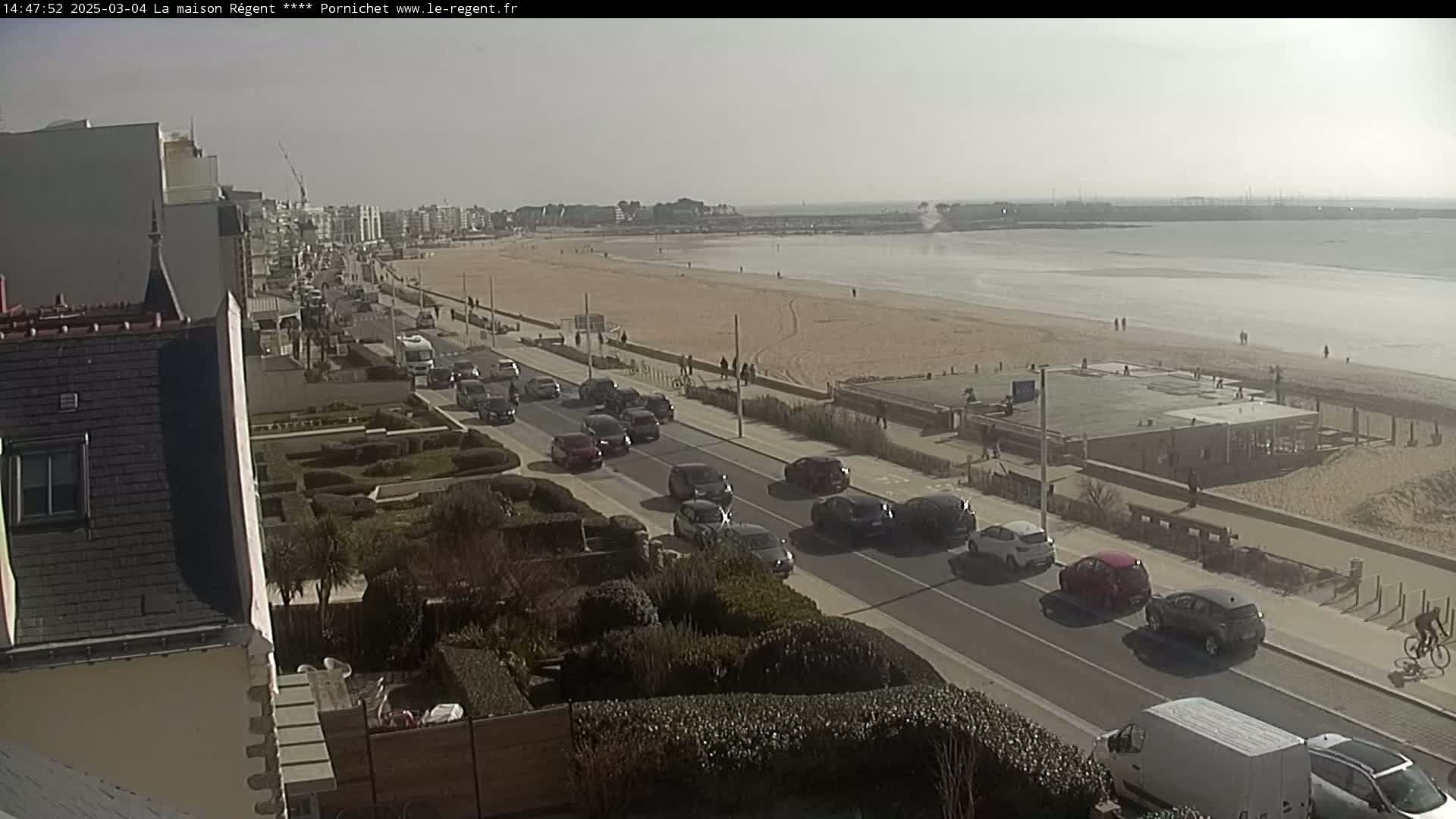 A sunny day reveals a beach with a few people, a road lined with cars, and buildings along a coastline.