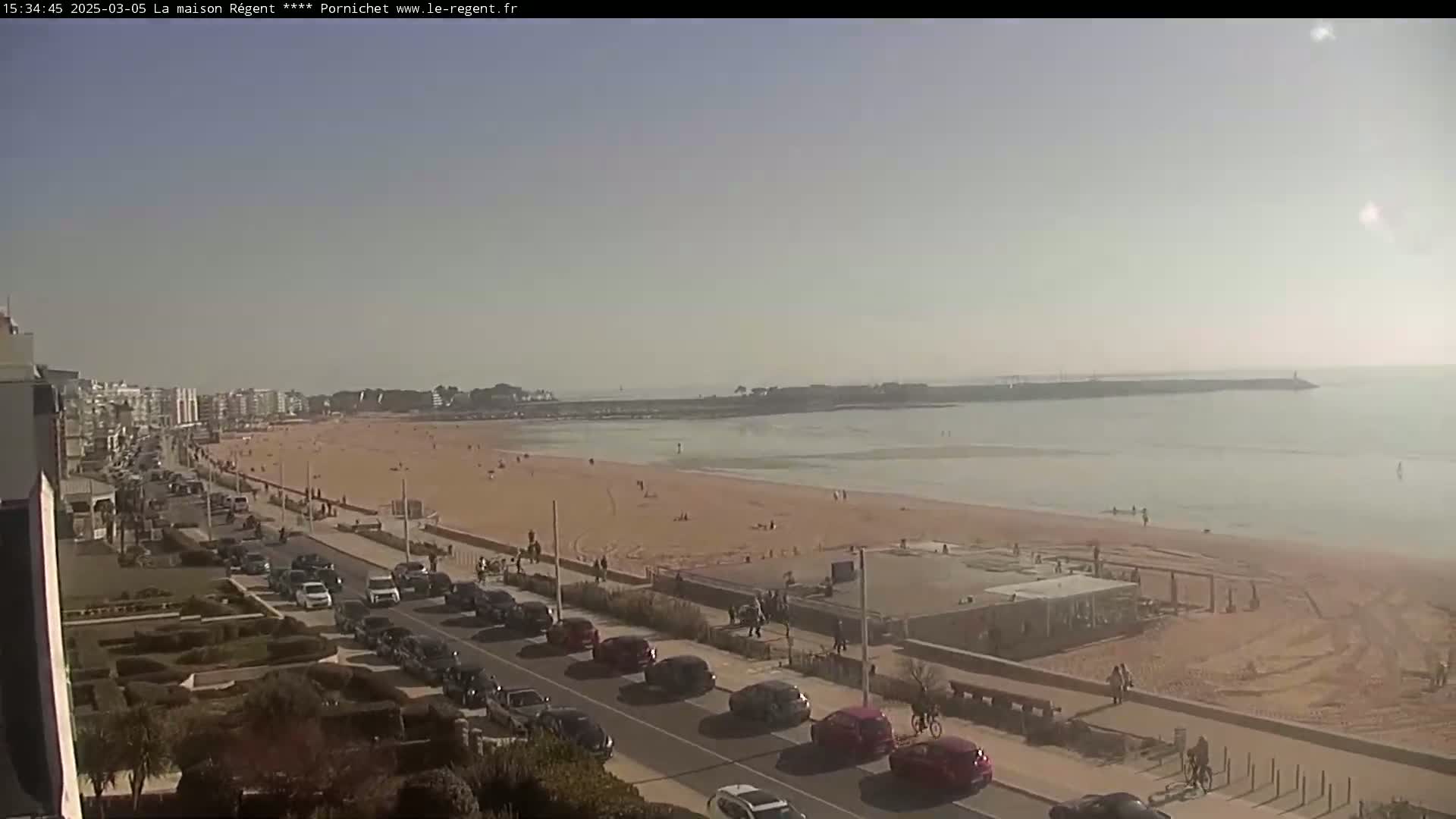 A sandy beach with a calm ocean under a clear, sunny sky is viewed from an elevated position, showing a road lined with parked cars and buildings in the background.