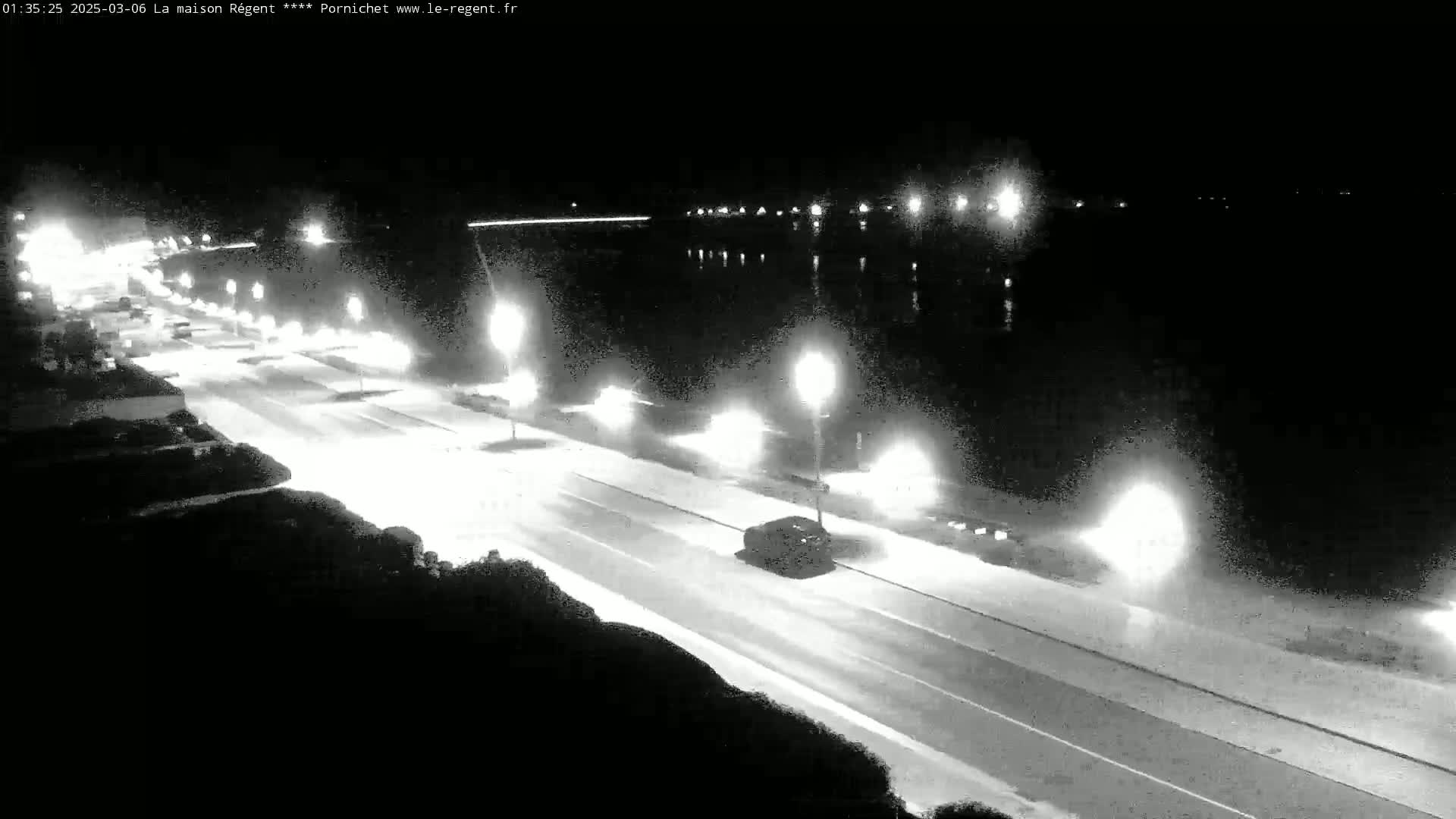 A nighttime, brightly lit road runs alongside a body of dark water, with vehicles traveling in both directions.