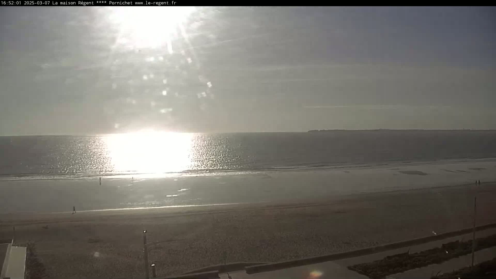 A mostly sunny day at a beach shows calm ocean waters reflecting sunlight, a wide sandy shore, and a few people walking on the sand.