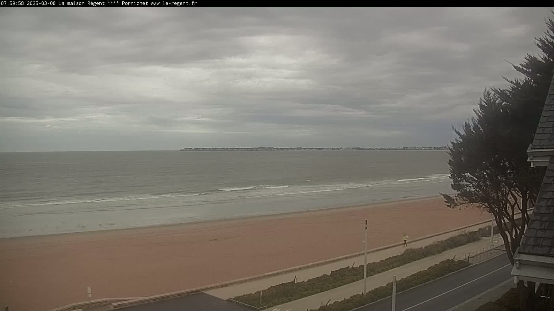 A sandy beach stretches along a calm, gray ocean under an overcast sky, with a distant shoreline visible and a person walking along the paved beachfront path.