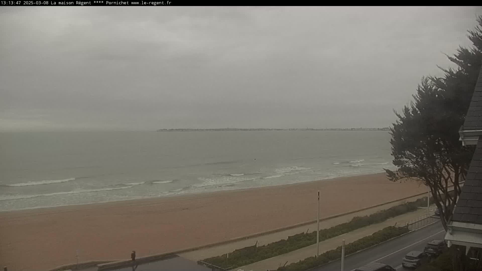 A wide shot of a beach on an overcast day shows calm waves lapping a sandy shore, with a road and vegetation visible in the foreground and a distant landmass on the horizon.