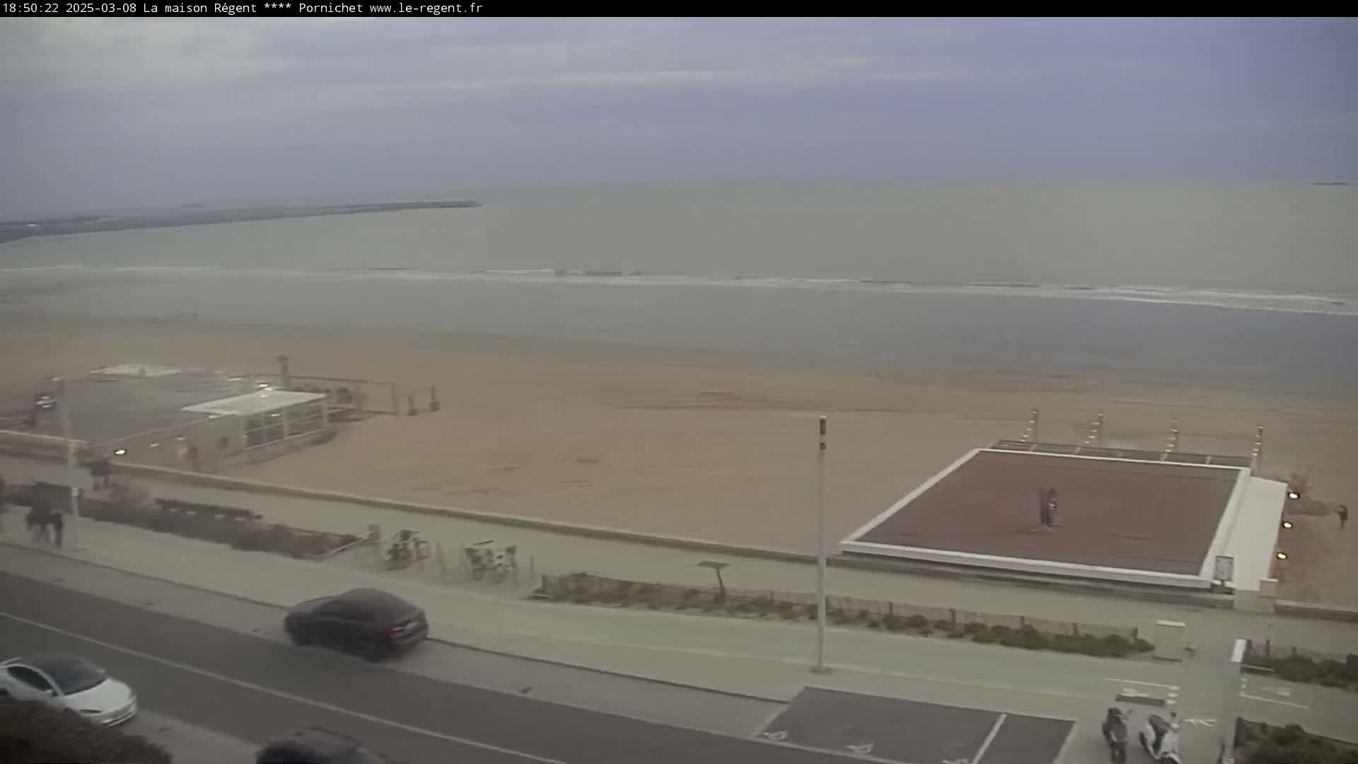 An overcast day at a beach shows a sandy shore with a beach volleyball court, some buildings, and cars parked along a paved road.