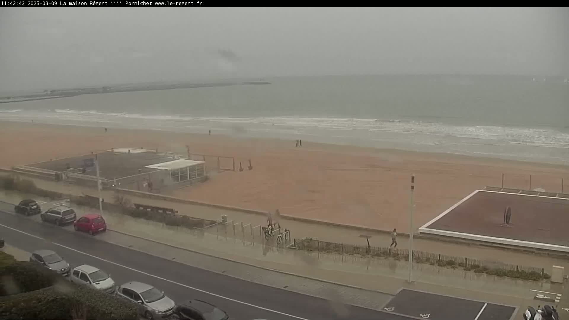 A wet, overcast beach scene shows a sandy beach with small waves, some people walking, and several cars parked along a road next to the beach.
