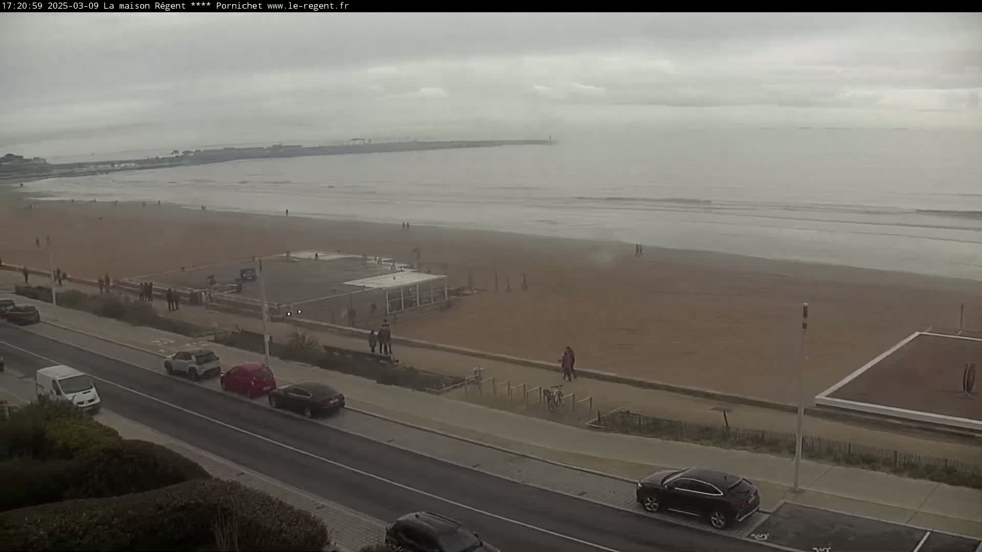 A wide shot shows a sandy beach with a few people and some small waves on an overcast day, next to a road with parked cars.
