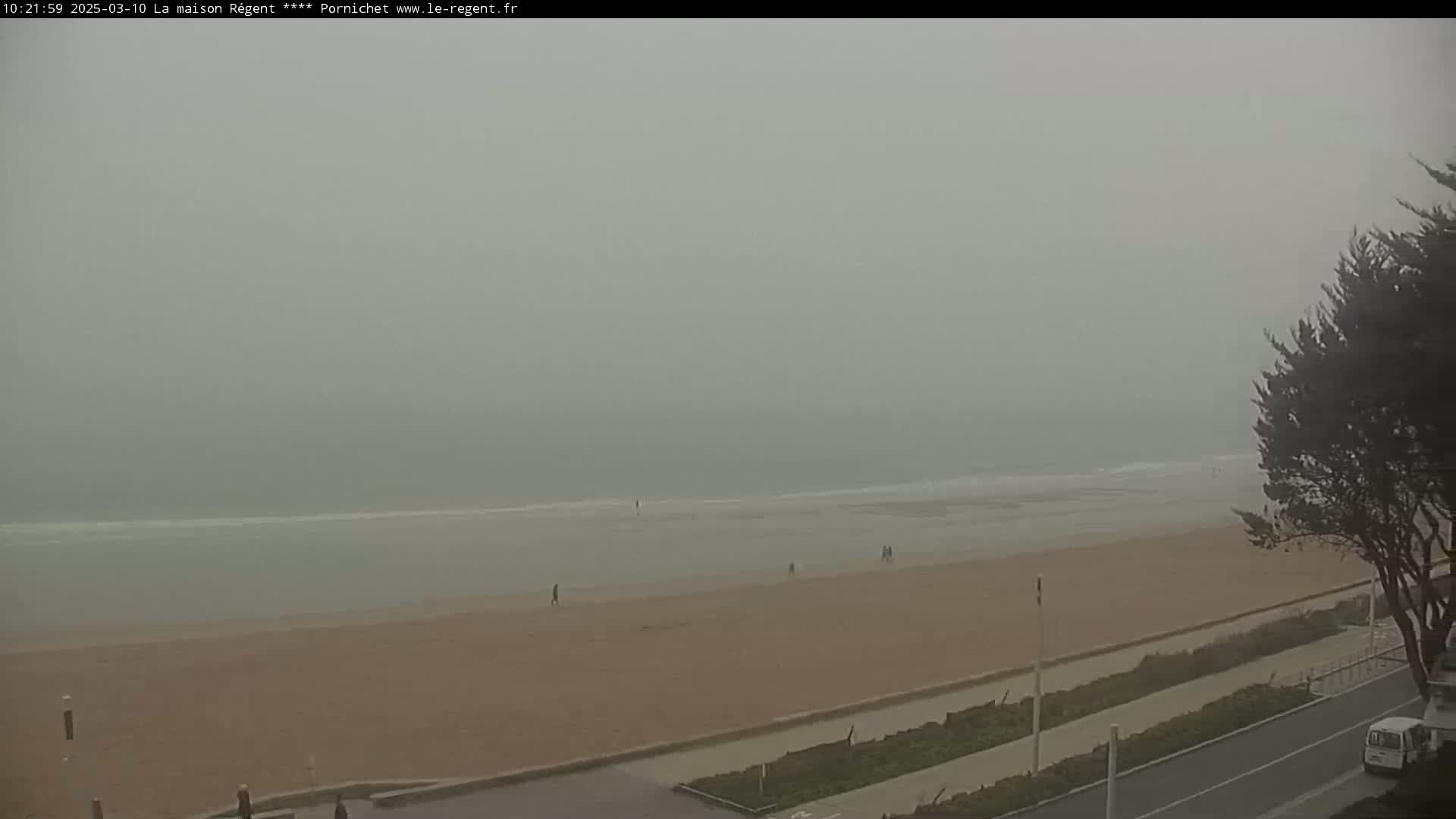 A hazy, foggy beach scene shows a wide sandy beach with a few people walking, a calm ocean, and vegetation along the road near the top right.
