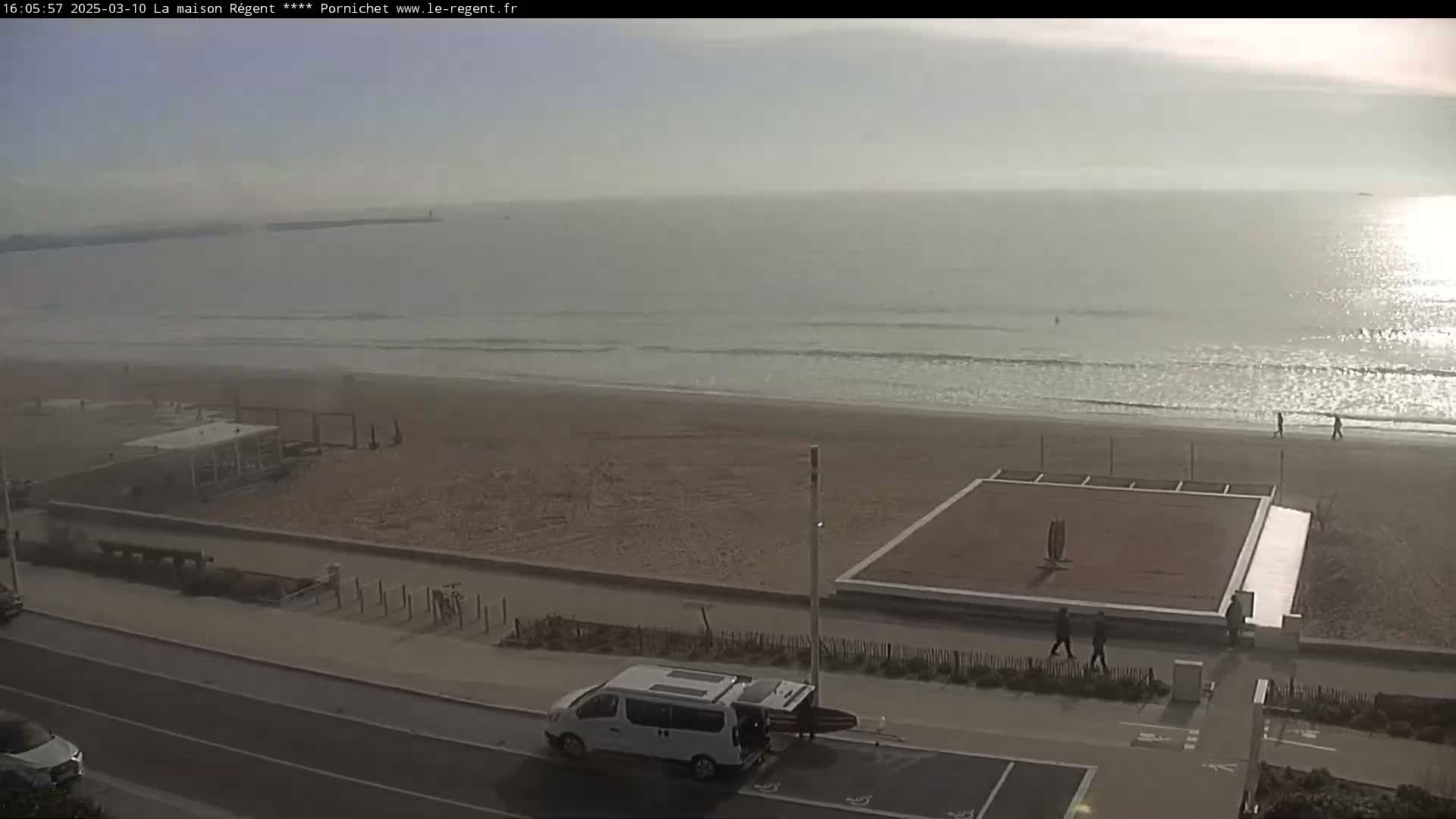 A sandy beach with a calm ocean under a hazy sky features a volleyball court and a white van parked near a road.