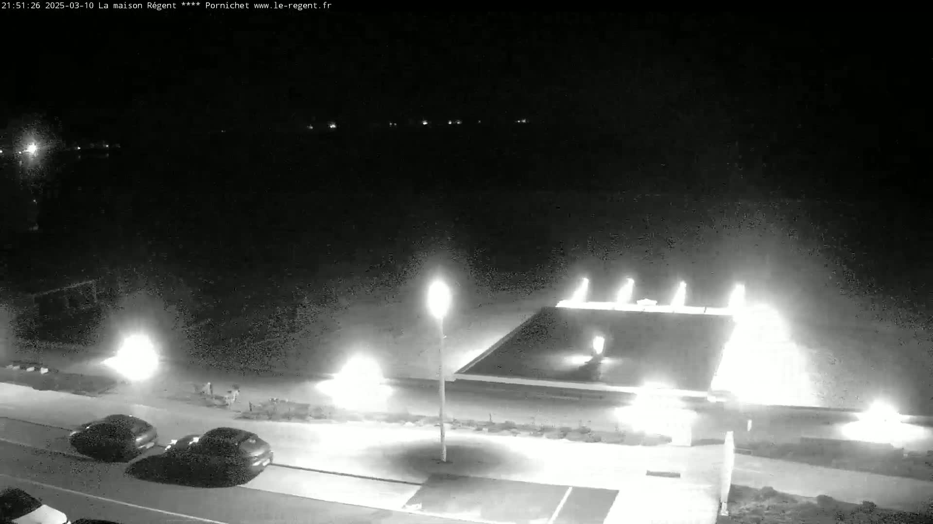 A nighttime, low-light view shows a paved area with parked cars near illuminated fountains and streetlights.