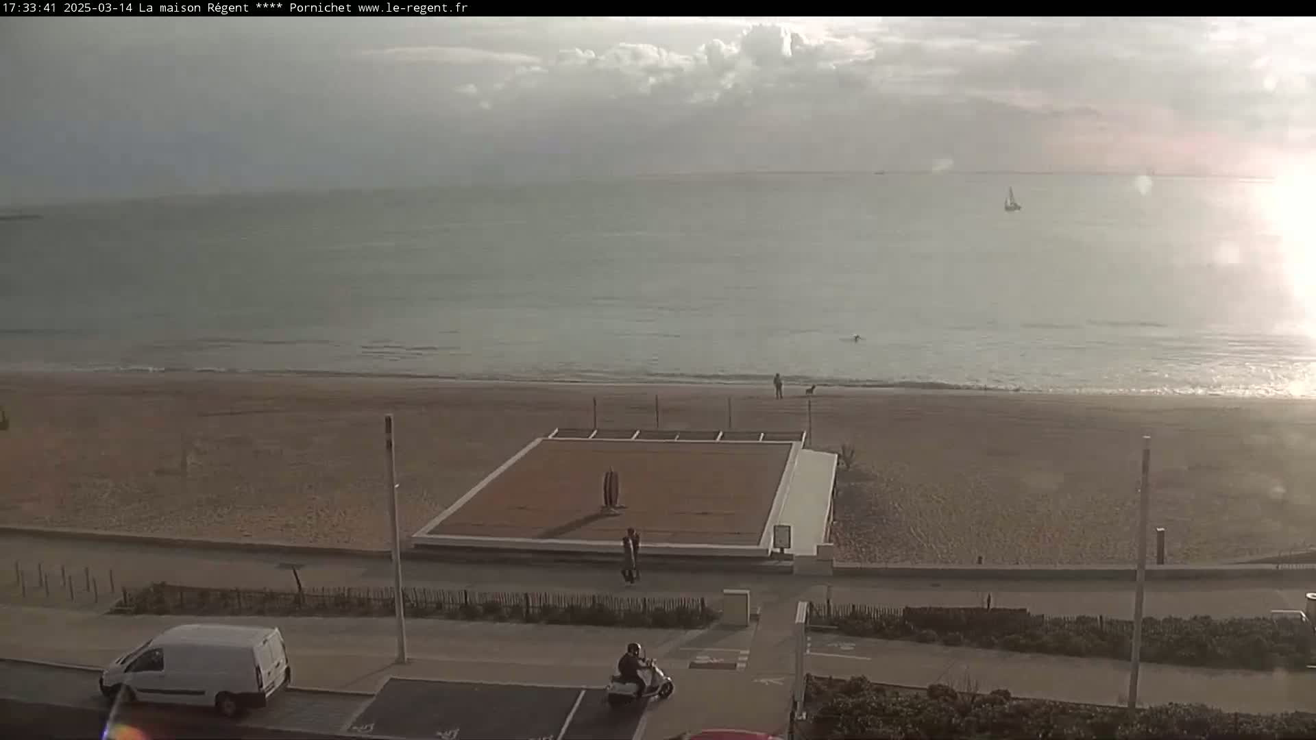 A mostly cloudy day shows a beach scene with a calm ocean, a sandy beach, a paved walkway, some people, vehicles, and a small structure on the sand.
