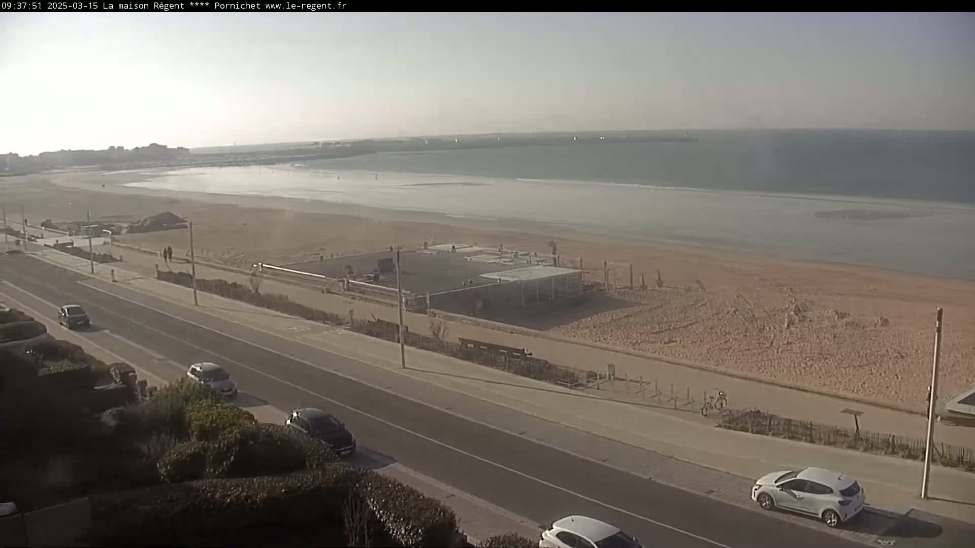 A long sandy beach stretches beside a road lined with cars under a hazy sky, with a calm ocean in the background.