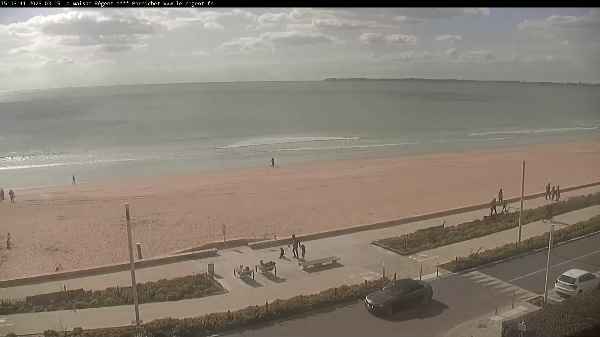 A mostly sunny day shows a wide sandy beach with a few people walking, a calm ocean, and a paved walkway with benches and a few cars parked nearby.