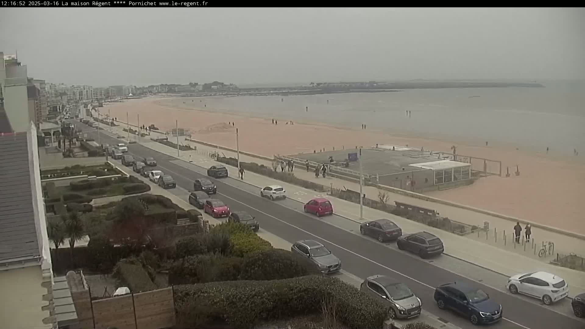 A seaside road with cars parked along it overlooks a wide, pink-sand beach with a few people strolling along the shore under an overcast sky.