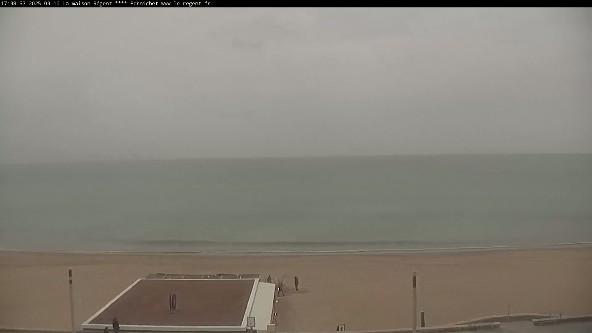 A mostly overcast sky hangs over a calm, light-green ocean meeting a sandy beach where a few people walk near a brown court.