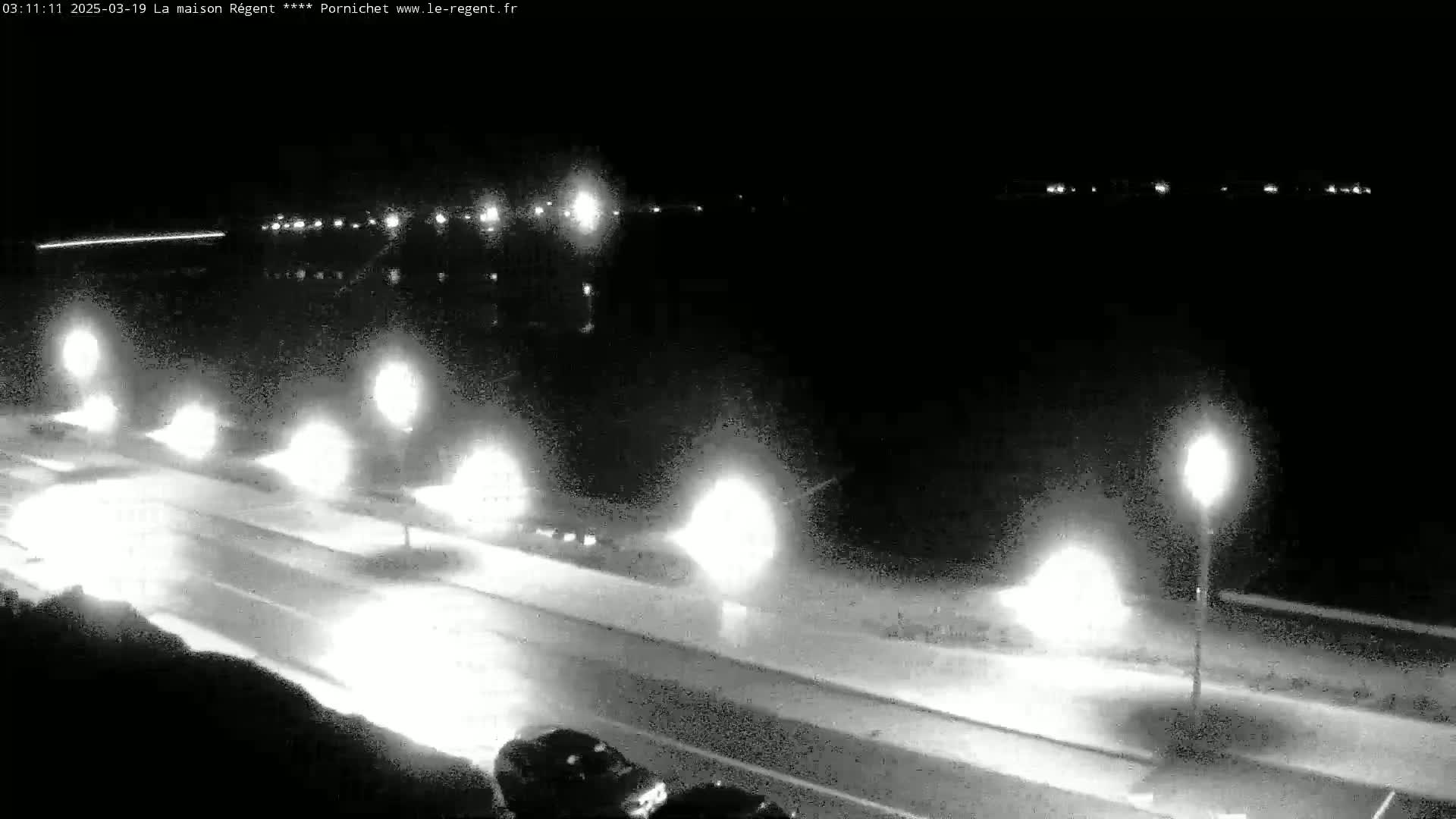 A nighttime, low-angle view shows a roadway with several vehicles driving under illuminated streetlights, beside a dark body of water.