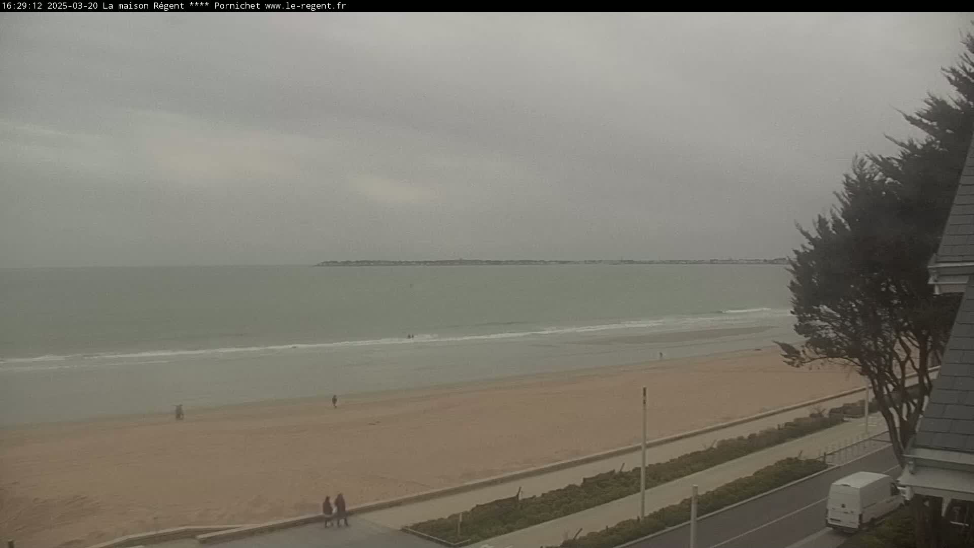 A wide shot of a sandy beach on an overcast day, with calm waves lapping the shore and a few people visible on the sand.