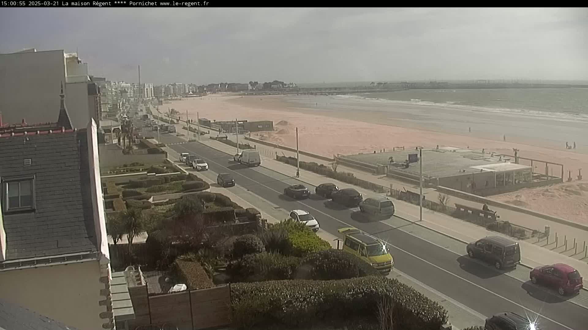 A seaside road with several cars is seen next to a wide, sandy beach under an overcast sky.