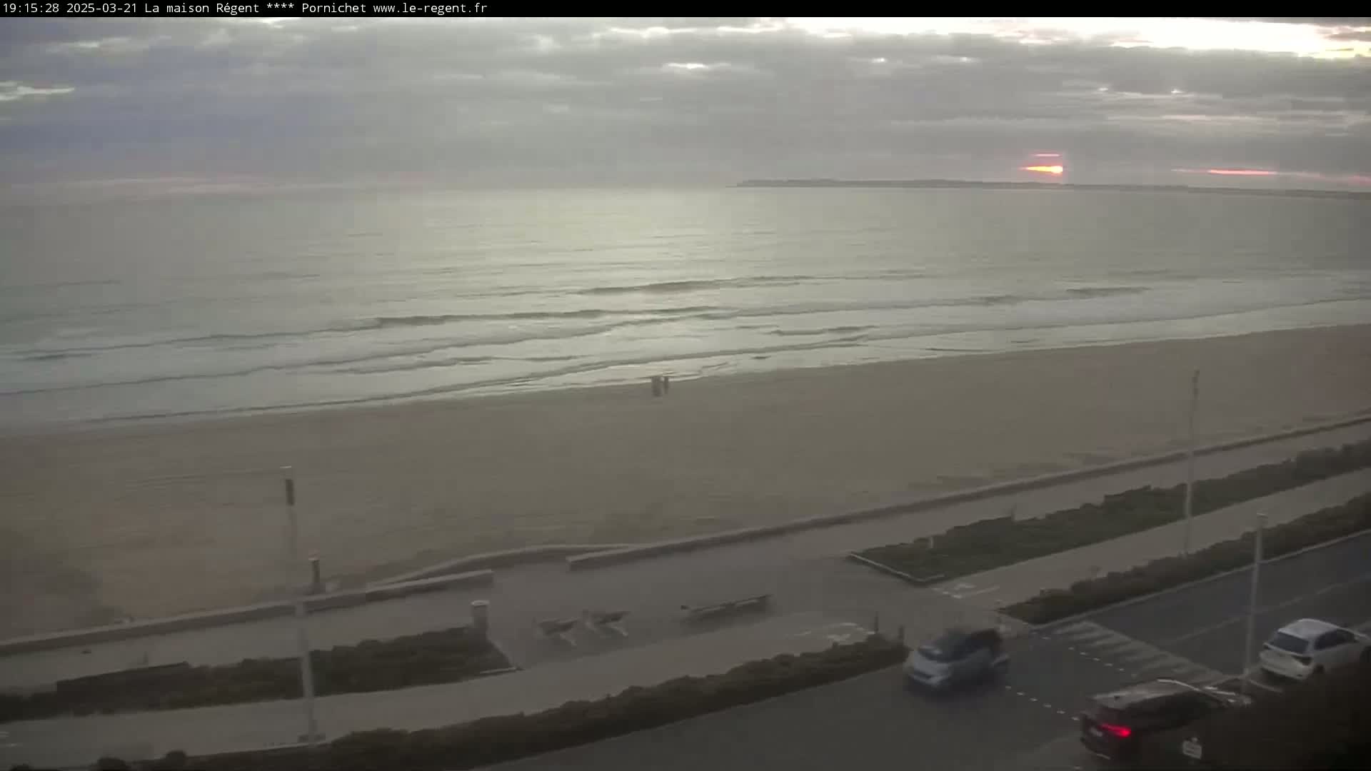 A mostly cloudy sunset over a calm ocean with small waves lapping a sandy beach, viewed from an elevated position overlooking a coastal road with several cars.