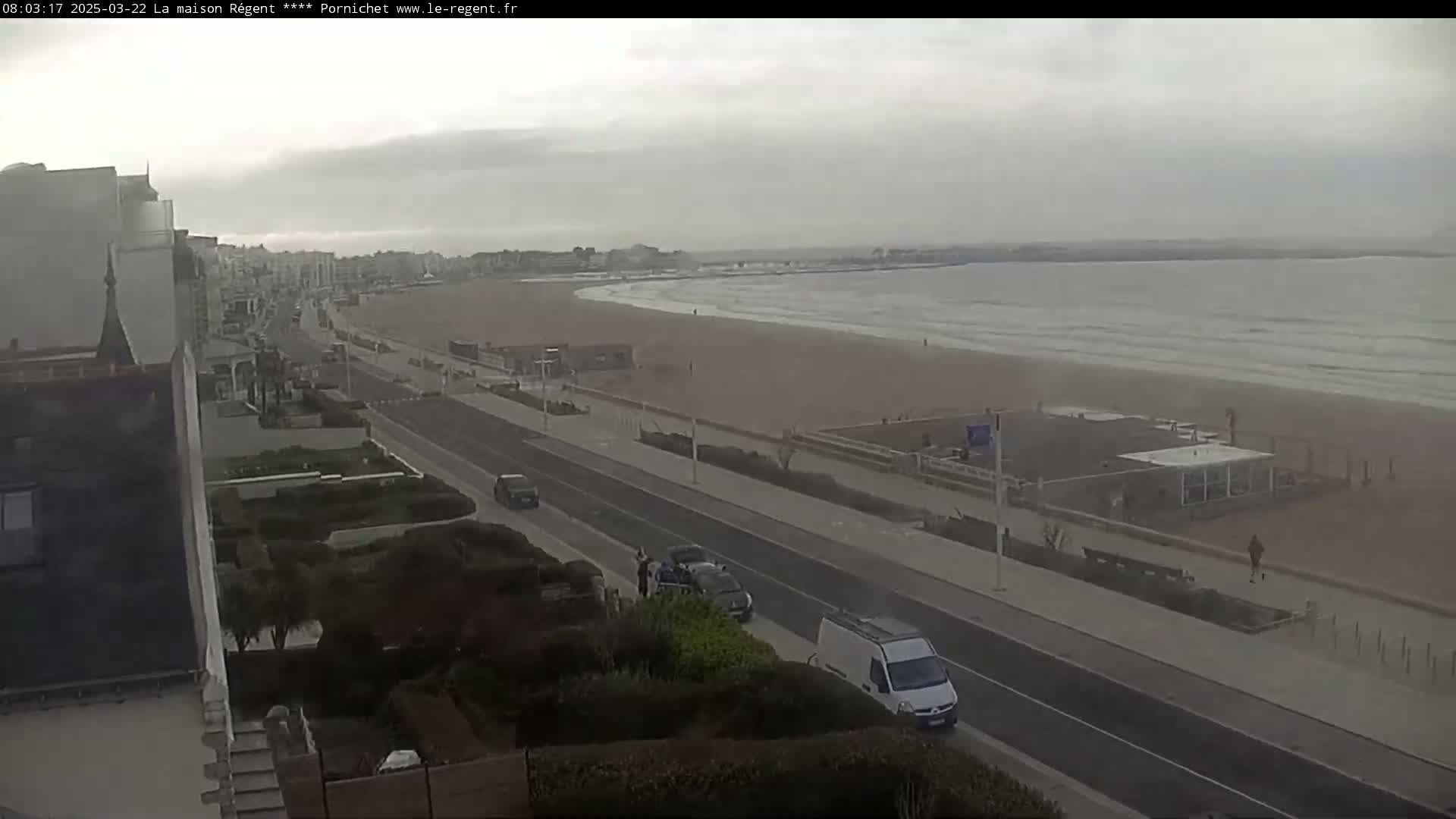 A long, sandy beach with gentle waves under an overcast sky is viewed from a high vantage point, showing a road and buildings along the coast.