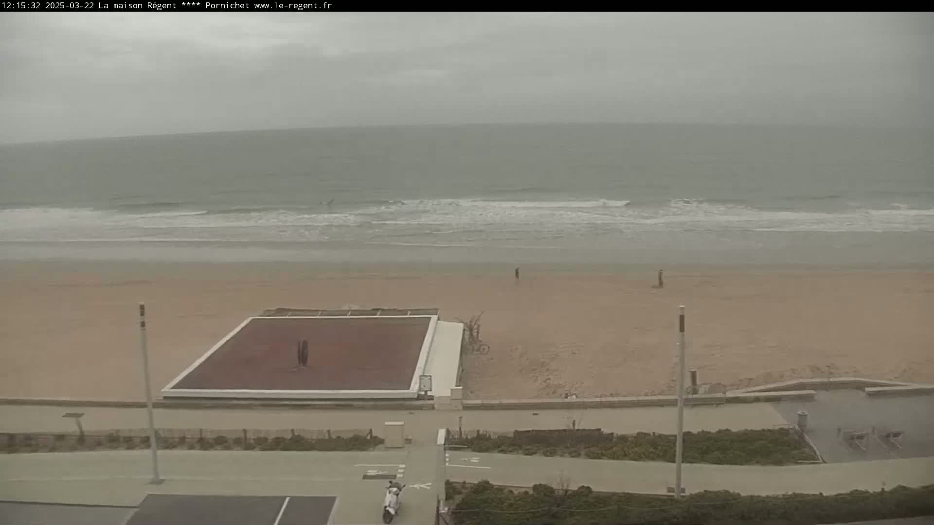 A sandy beach with a small, dark-colored court is visible under an overcast sky, with gentle waves rolling onto the shore.