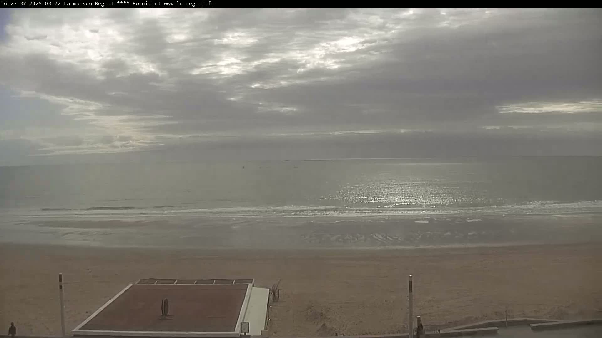 A mostly cloudy sky overlooks a calm ocean meeting a sandy beach with a flat-roofed structure on the shore.