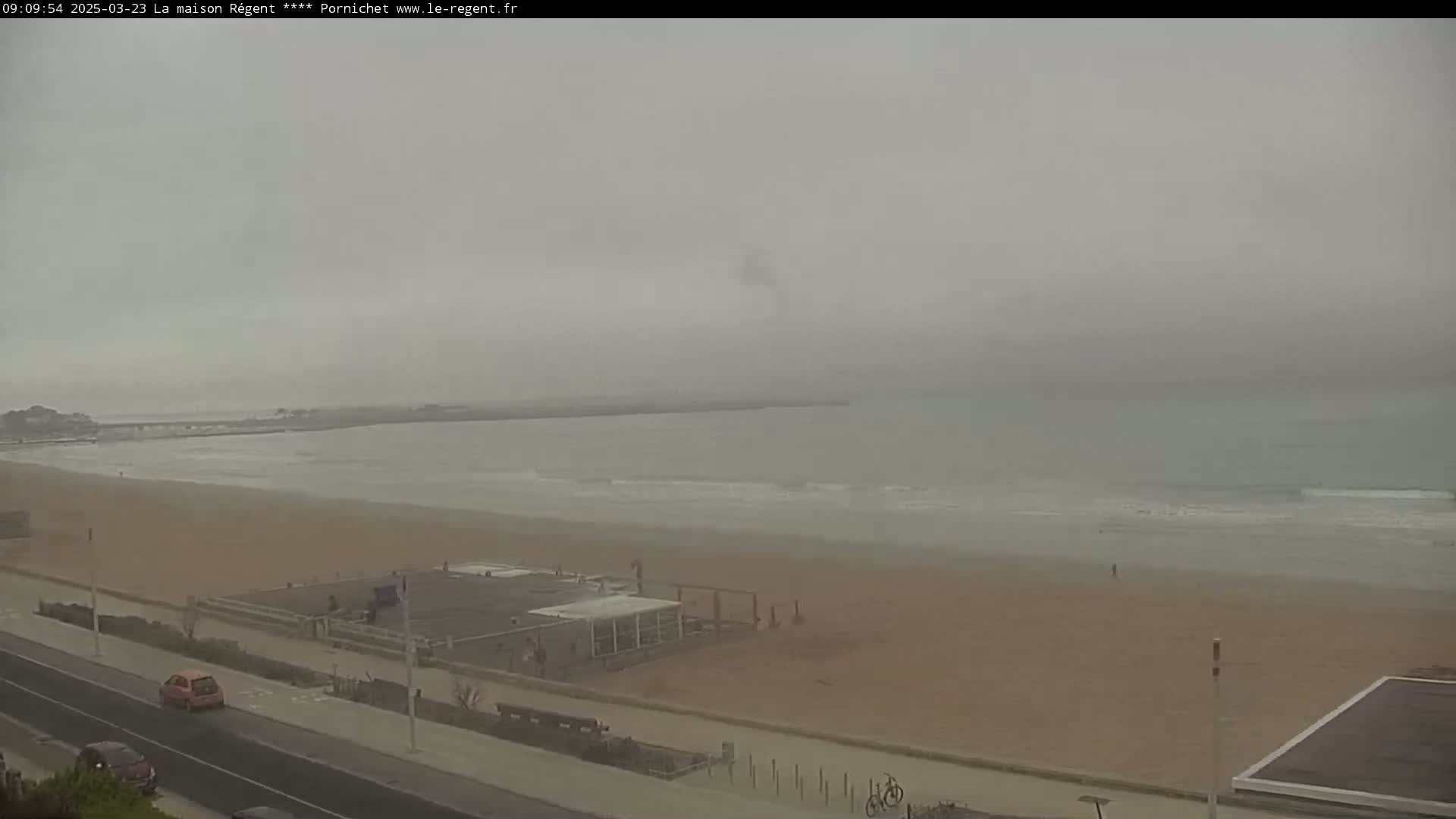 A foggy view of a beach with calm waves, a road, and a few buildings.