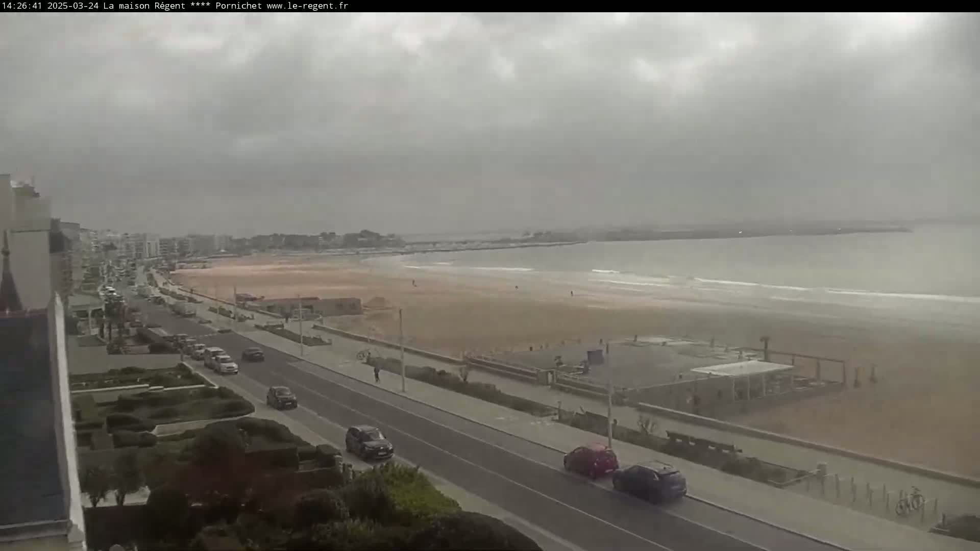A long, sandy beach with a few people and cars on a road that runs alongside it, under an overcast sky.