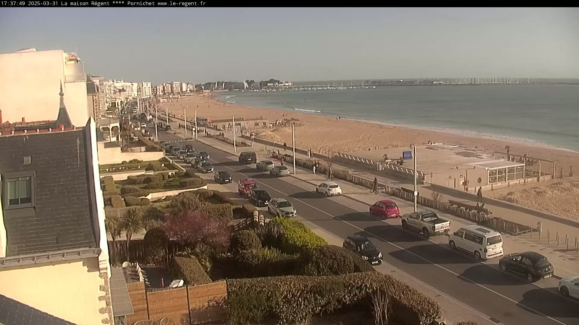 A sunny day at a beach with a sandy shore, calm ocean water, and a road lined with cars and buildings.