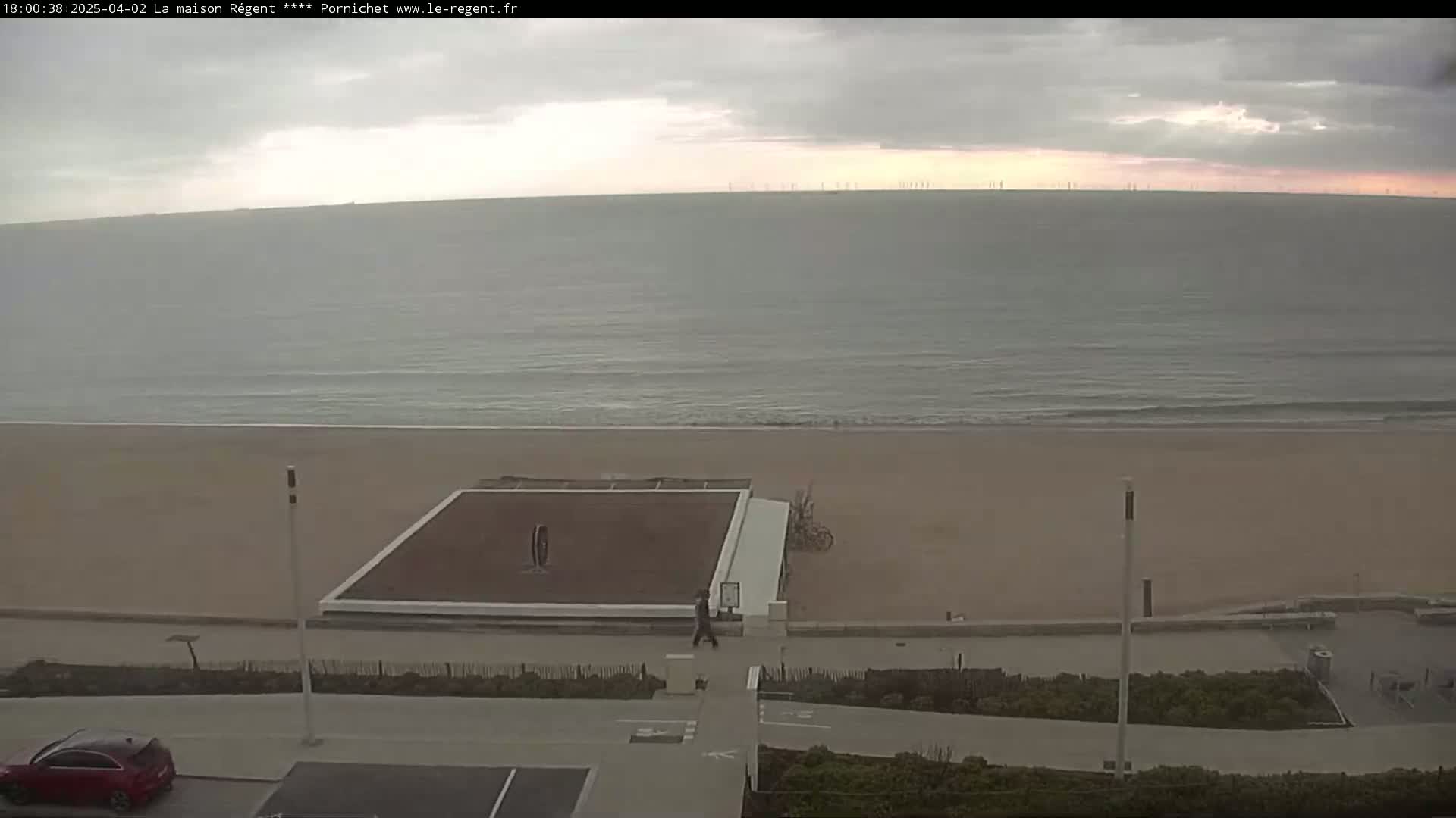 A beach with a calm ocean under an overcast sky, featuring a paved area with a small court and a person walking.